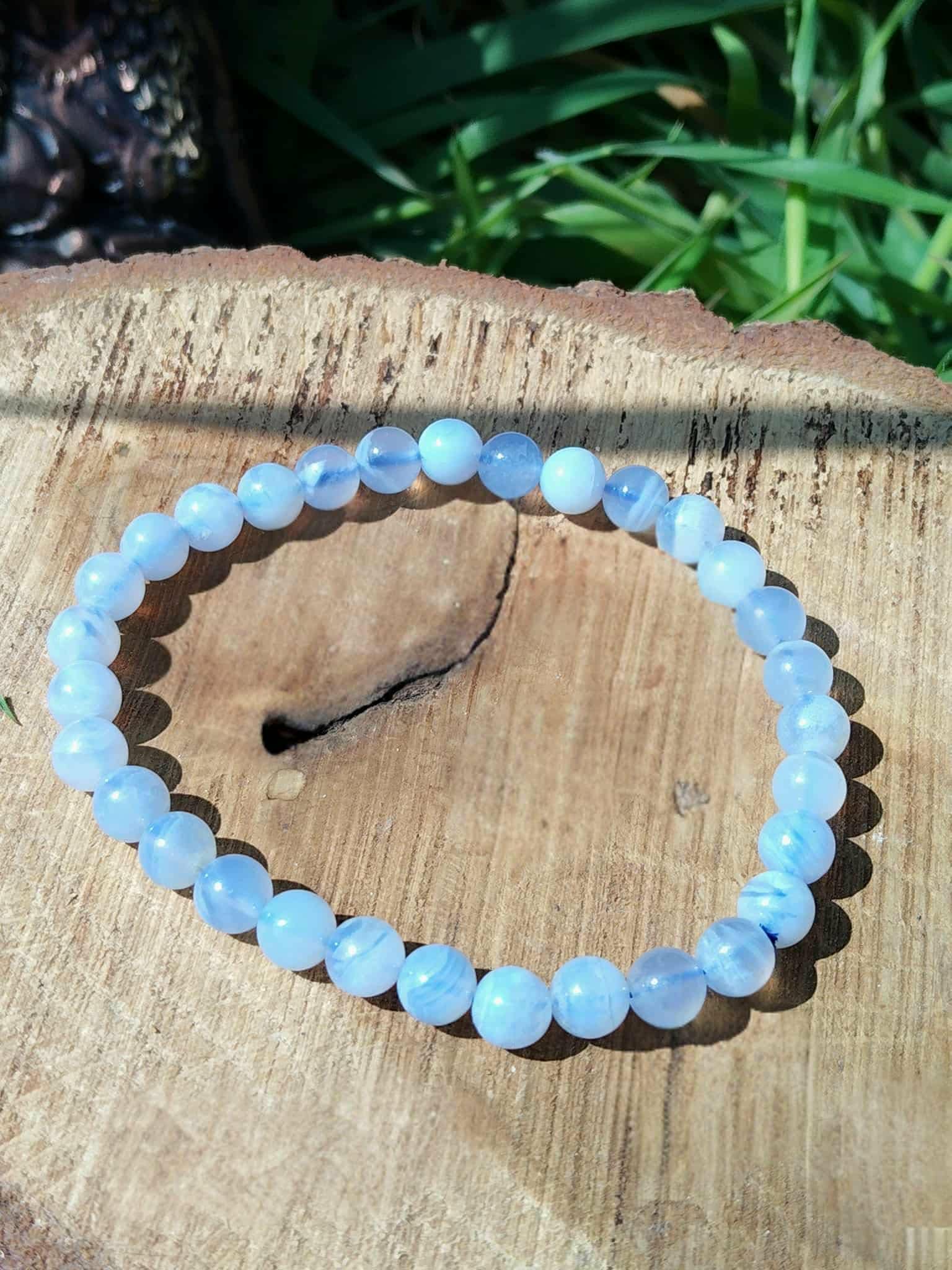 Bracelet calcédoine bleue 6 mm sur tranche de bois clair veinée avec herbe floue, parole douce, calme du cœur et fluidité émotionnelle.