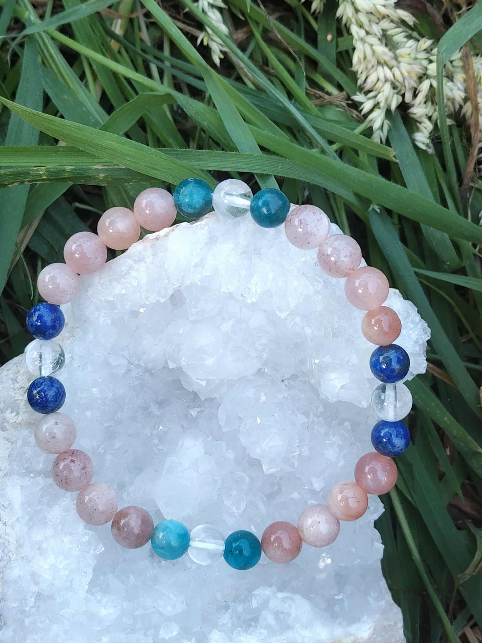 Bracelet bien-être 6 mm posé sur géode de quartz blanc dans l’herbe, lapis-lazuli, pierre de soleil, apatite et cristal de roche pour clarté mentale et parole apaisée