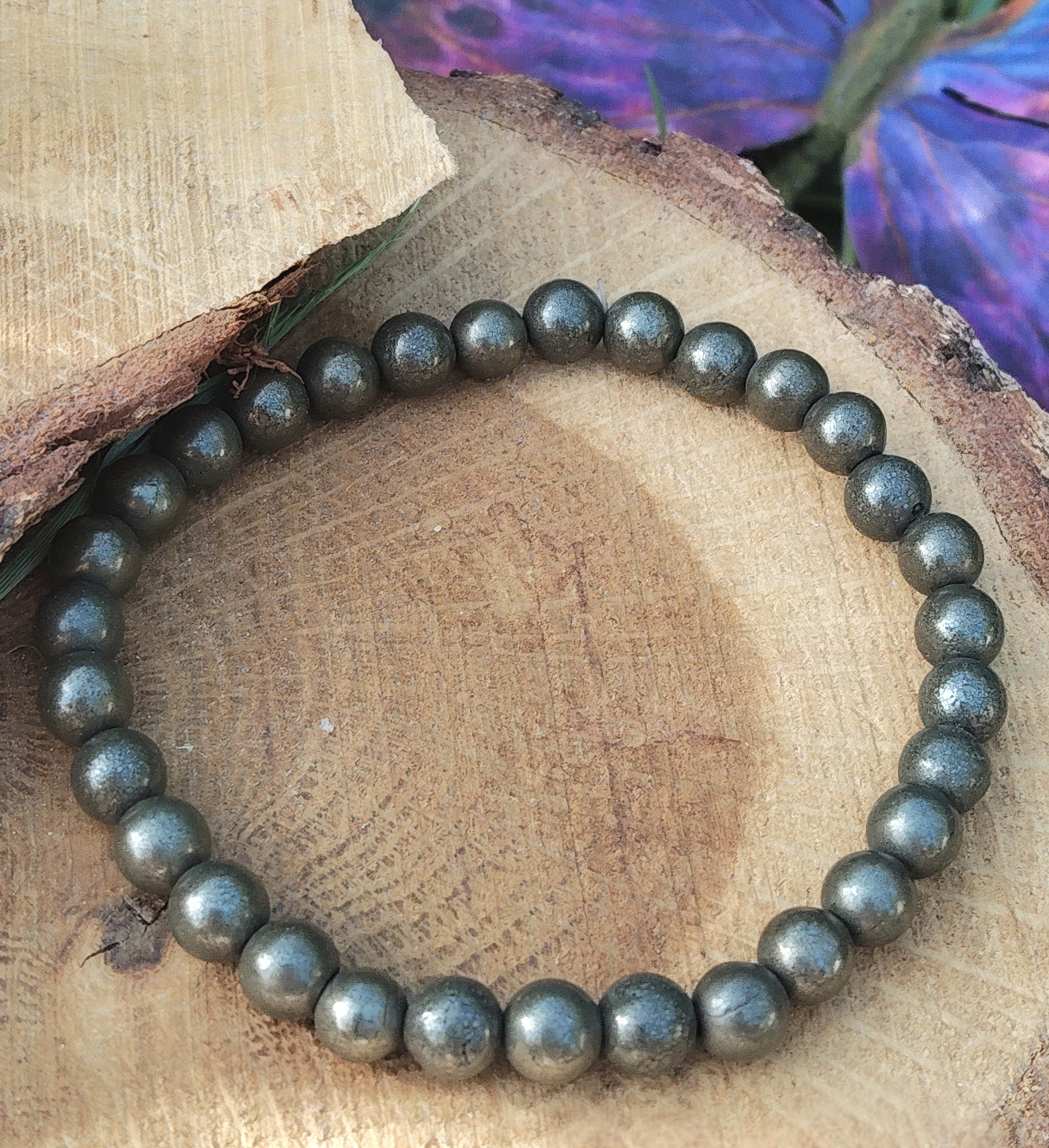 Bracelet pyrite 6 mm gris anthracite métallisé en perles rondes posé sur un rondin de bois, avec aile violette en décor, évoquant la maîtrise de soi, l’élan protecteur et la solidité intérieure
