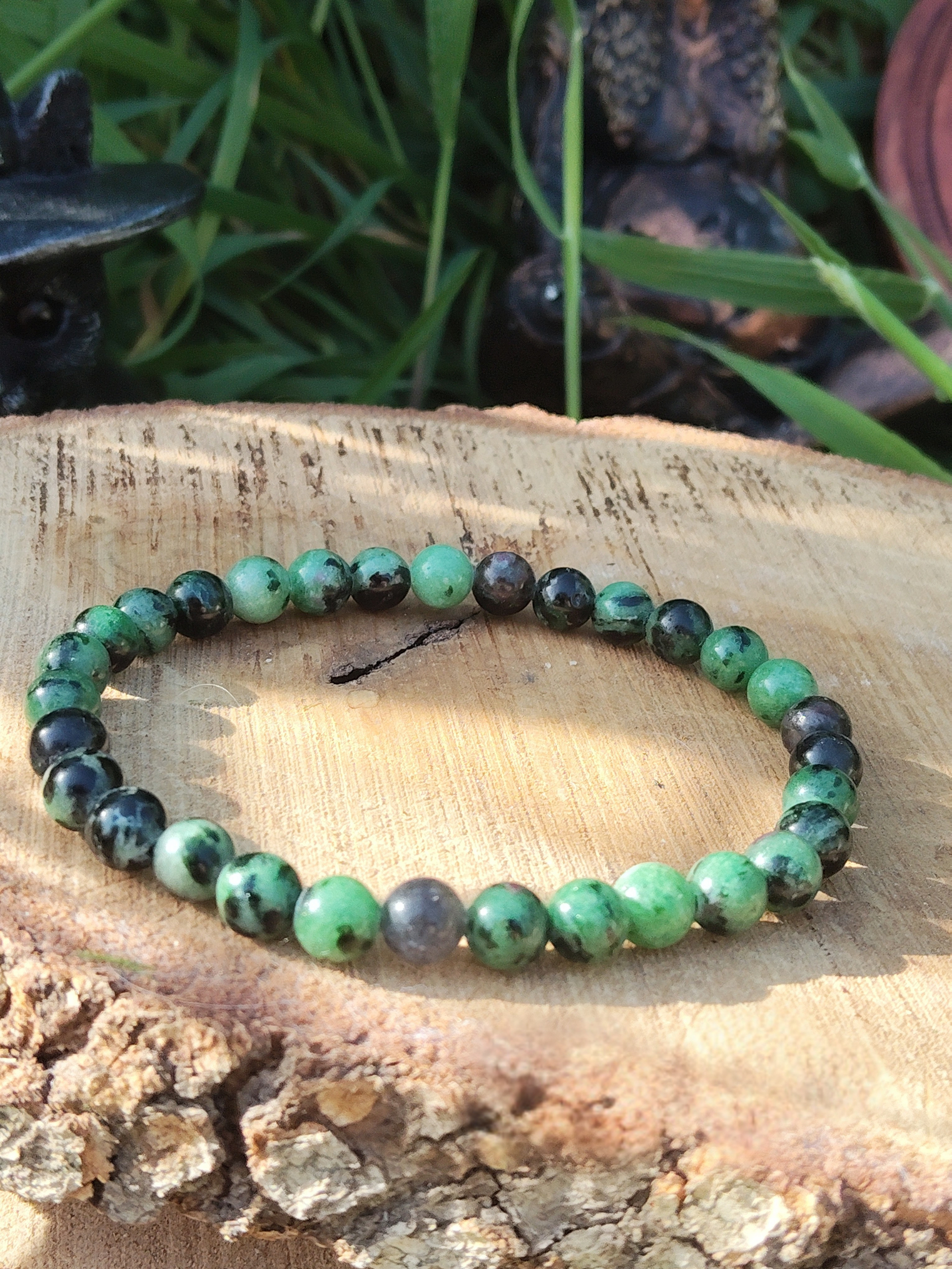 Bracelet rubis zoïsite 6 mm sur tranche de bois avec écorce, herbe floutée et petites statuettes, ancrage stable, courage tranquille et confiance en soi