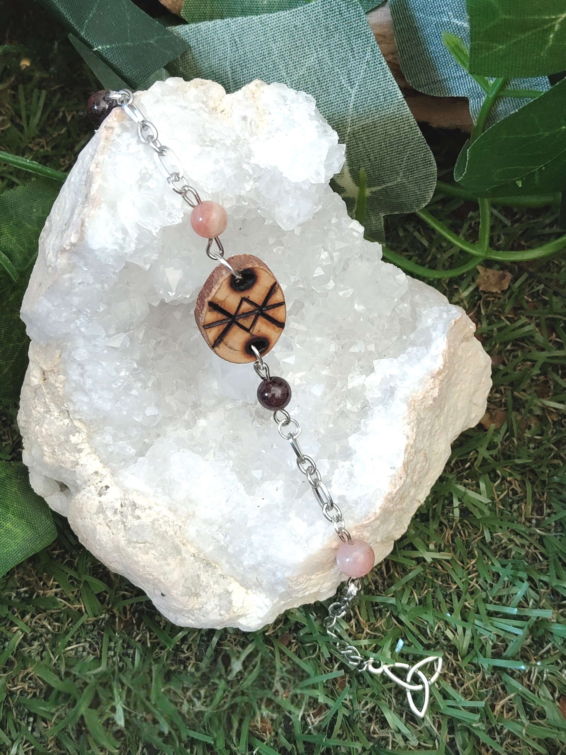 Bracelet rune de protection en bois création Magic Gaïa sur chaîne argentée, perles rose pâle et bordeaux, posé sur amas de quartz blanc avec feuilles vertes sur herbe; protection douce, ancrage, sérénité.