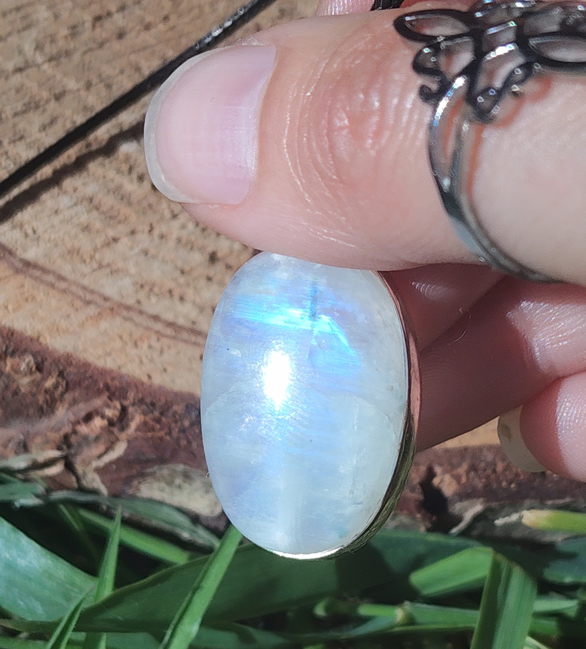 Pendentif labradorite blanche ovale en argent 925, tenu entre les doigts au soleil, fond bois et herbe, pierre naturelle évoquant le recentrage, l’apaisement et la sensibilité intuitive.