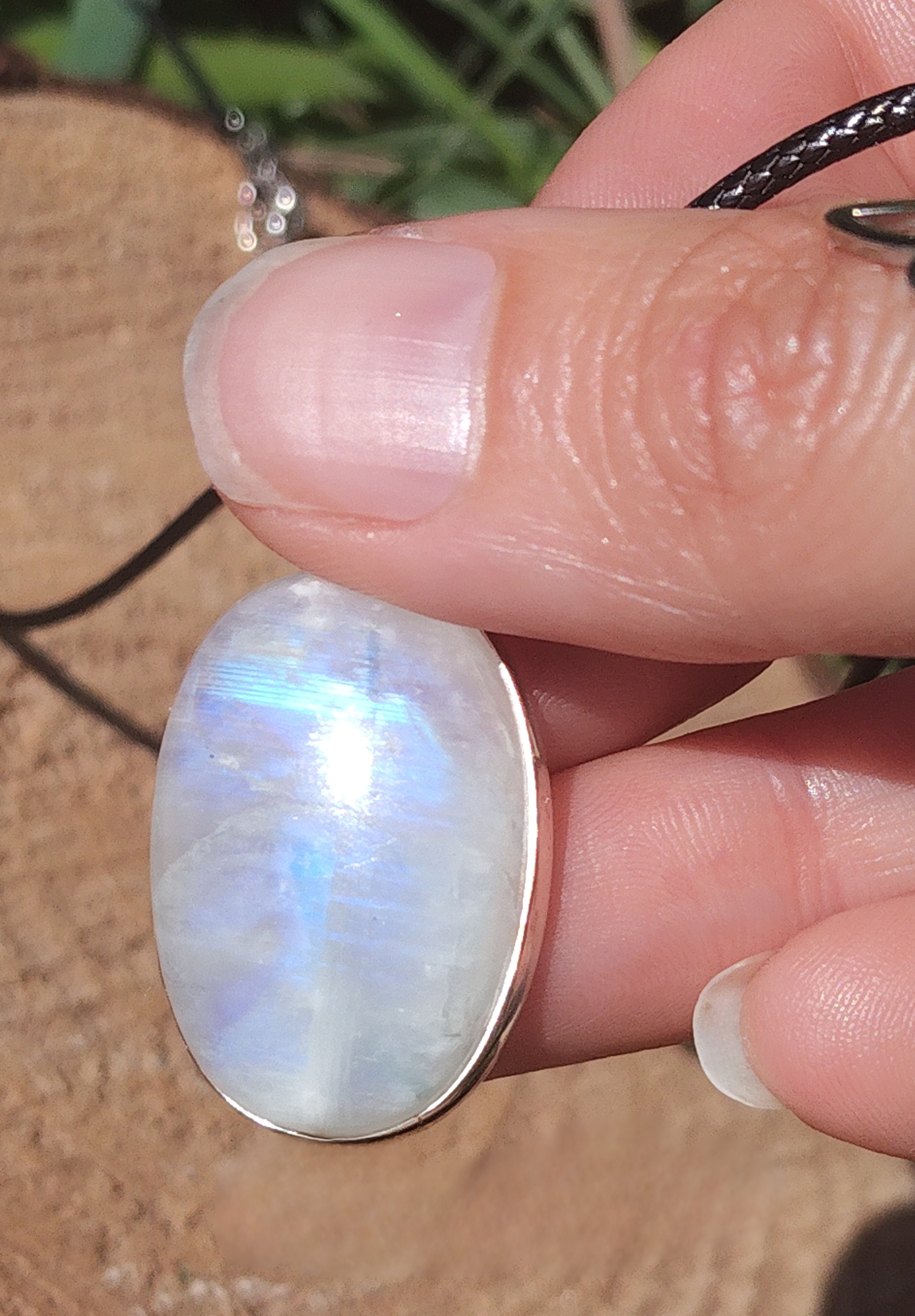 Pendentif ovale en labradorite blanche argent 925, tenu entre les doigts au soleil avec reflet bleu, cordon noir visible, bijou en pierre naturelle associé à la délicatesse, au calme et à l’inspiration intérieure.