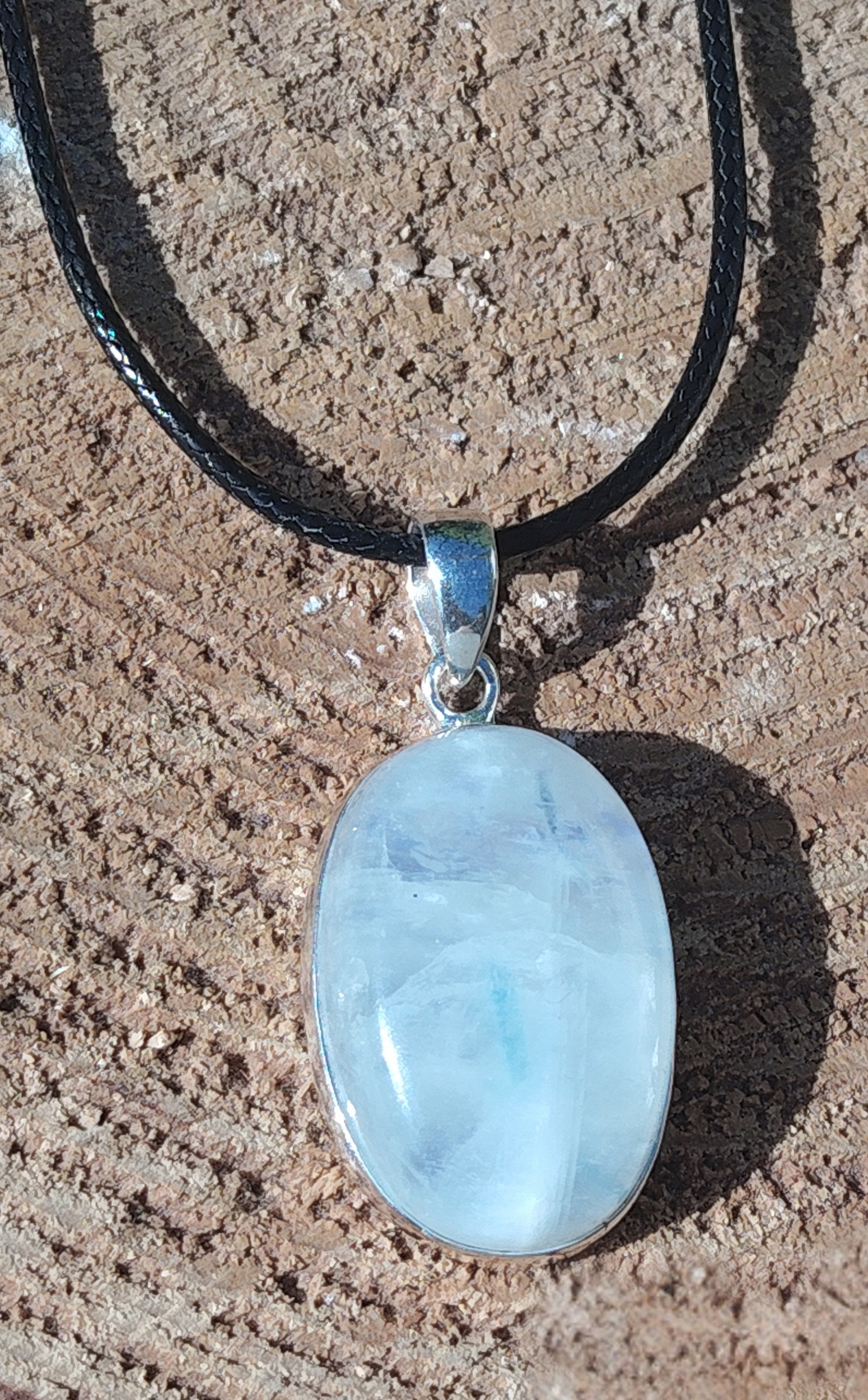 Pendentif ovale en labradorite blanche sertie argent 925 sur cordon noir, posé sur pierre beige au soleil, bijou en pierre naturelle lié à la clarté d’esprit, à la tranquillité et à la finesse intuitive.