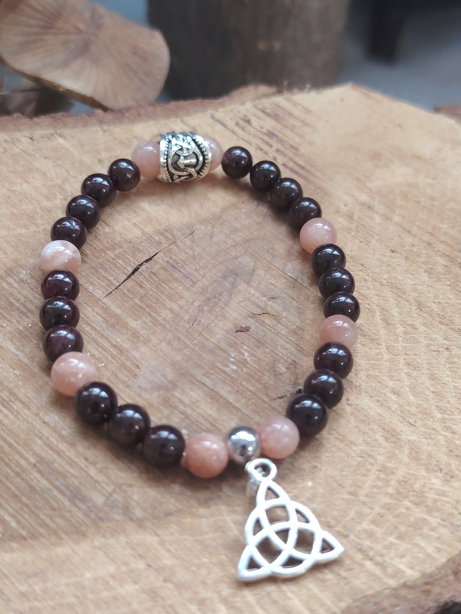 Bracelet viking en pierre de soleil et grenat posé sur bois clair, triquetra argentée et rune centrale, détermination, stabilité et esprit guerrier