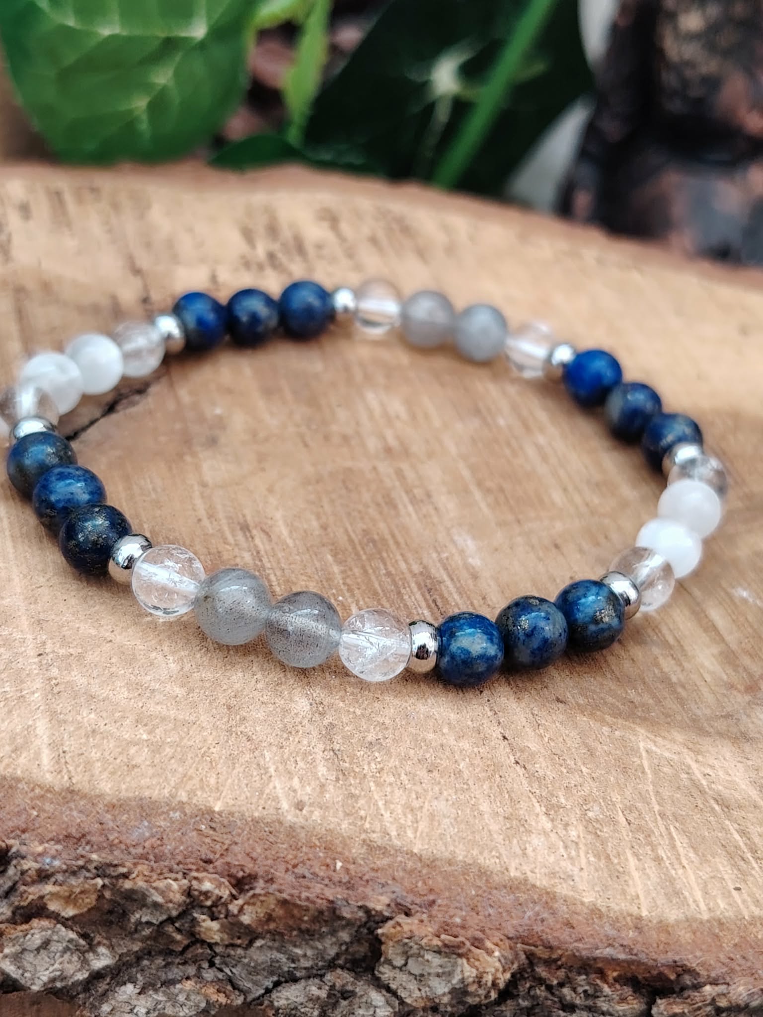 Bracelet migraine 6 mm en lapis lazuli, labradorite et sélénite présenté sur un rondin de bois, avec feuillage sombre en arrière-plan, pour clarté, apaisement et détente.