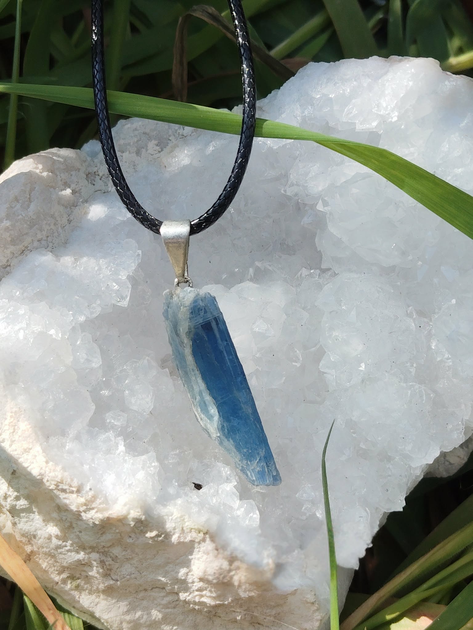 Pendentif cyanite bleue brute sur cordon noir, bélière argentée, posé sur amas de quartz blanc et herbes; intention: apaisement, expression.