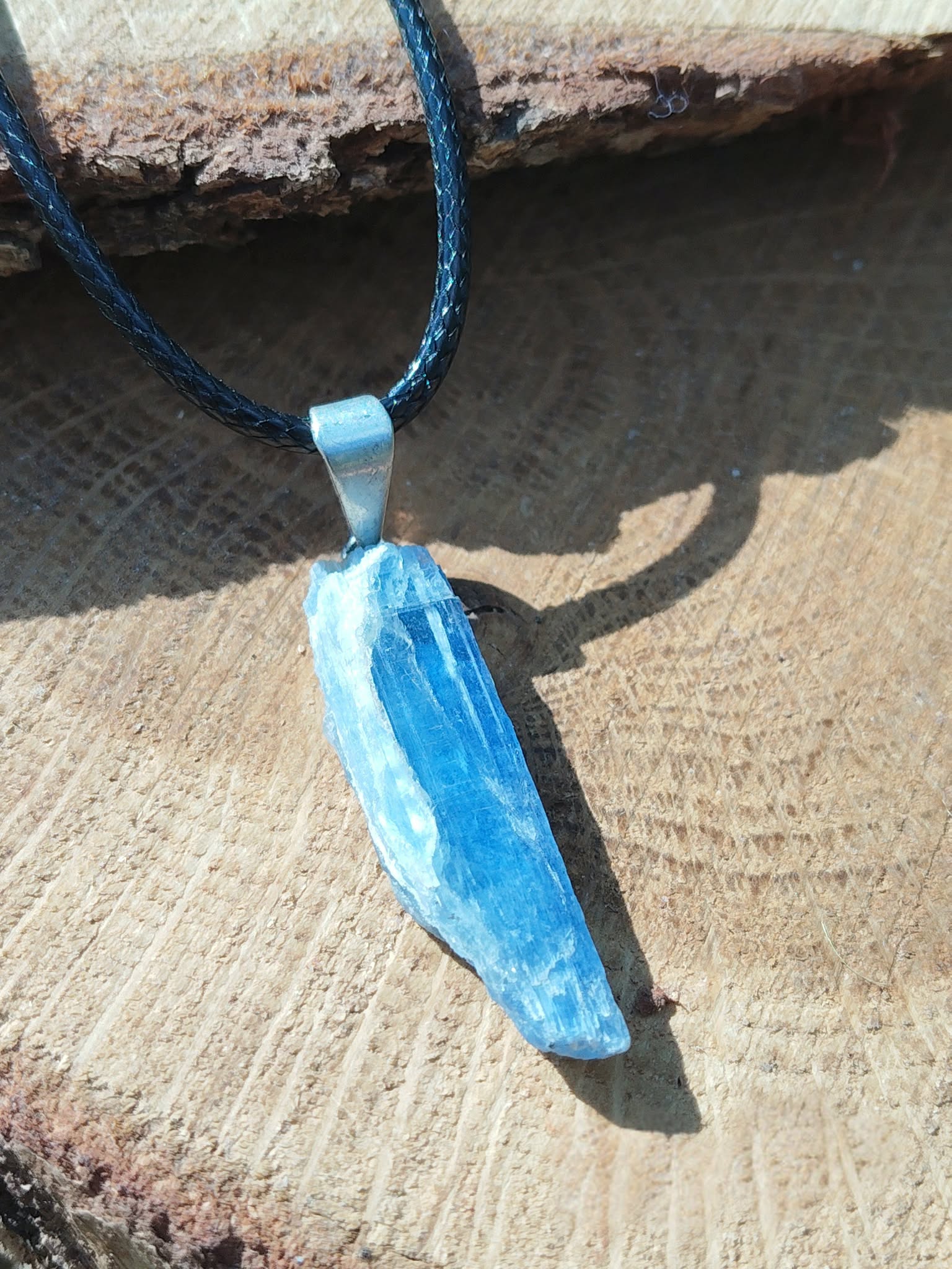 Pendentif en cyanite bleue brute sur cordon noir, bélière argentée, posé sur rondin de bois au soleil; intention: clarté, apaisement.