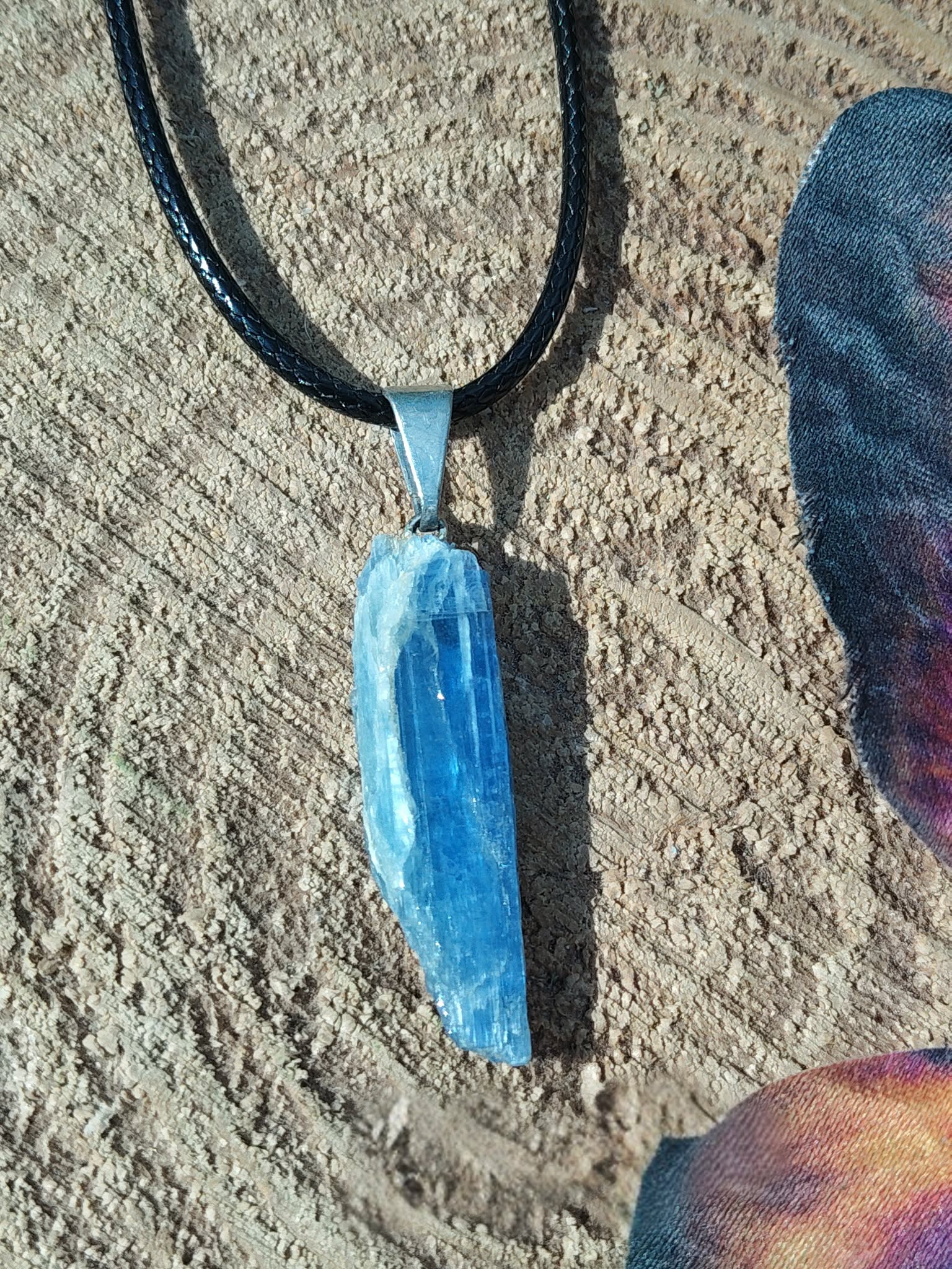 Pendentif cyanite bleue brute sur cordon noir, bélière argentée, posé sur bois et ailes de papillon; intention: clarté, sérénité.