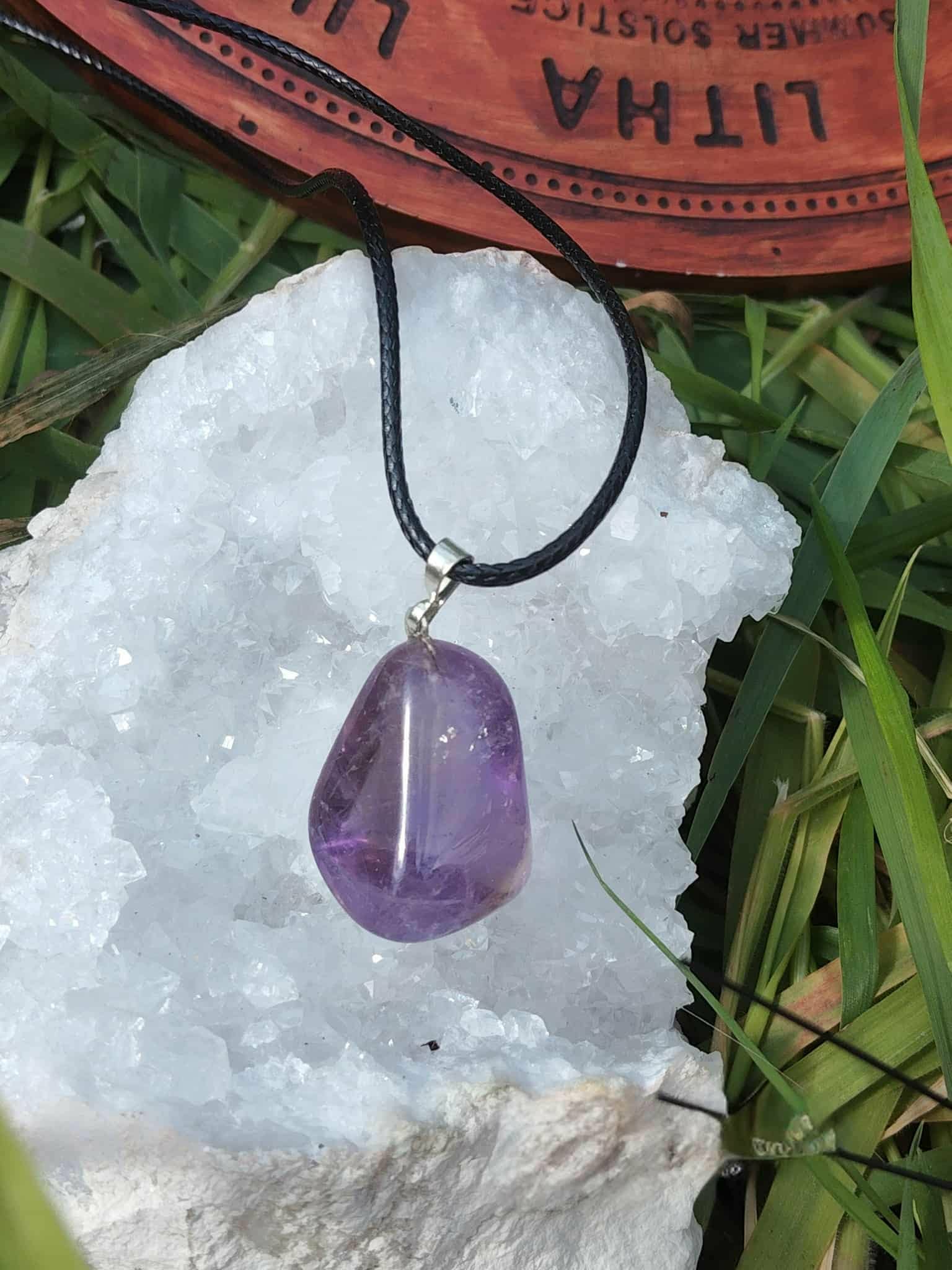 Pendentif en amétrine violette sur cordon noir, présenté sur un amas de quartz  et décor végétal, pierre naturelle associée à la paix intérieure, à la finesse d’esprit et à l’équilibre émotionnel.
