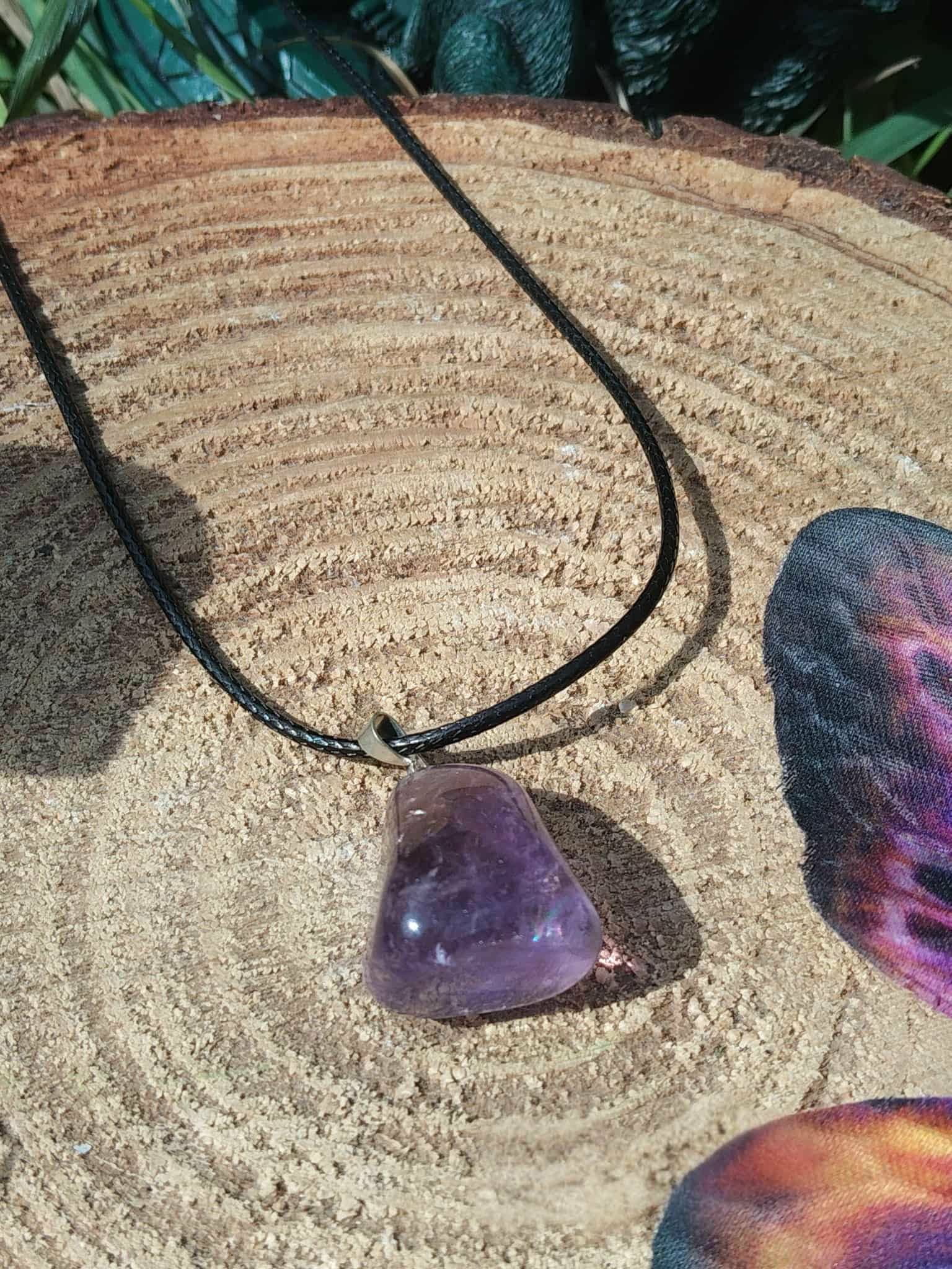 Pendentif en amétrine violette sur cordon noir, posé sur bois avec décor minéral en fond, pierre naturelle reliée au discernement, au recentrage et à la sérénité émotionnelle.