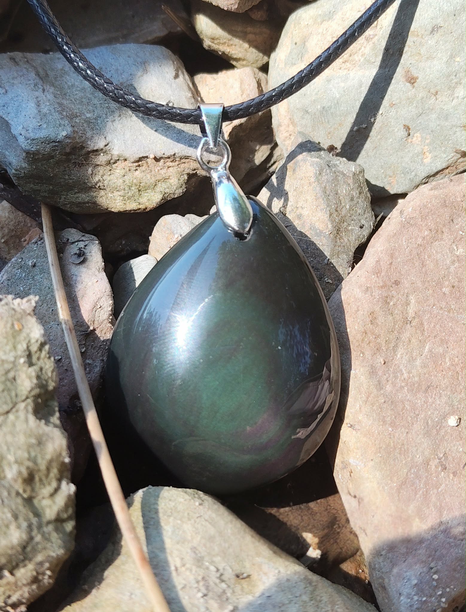 Pendentif obsidienne œil céleste en goutte sur cordon noir, posé entre galets clairs au soleil avec reflets verts et pourpres, bijou en pierre naturelle évoquant la force intérieure, l’enracinement et la vigilance.