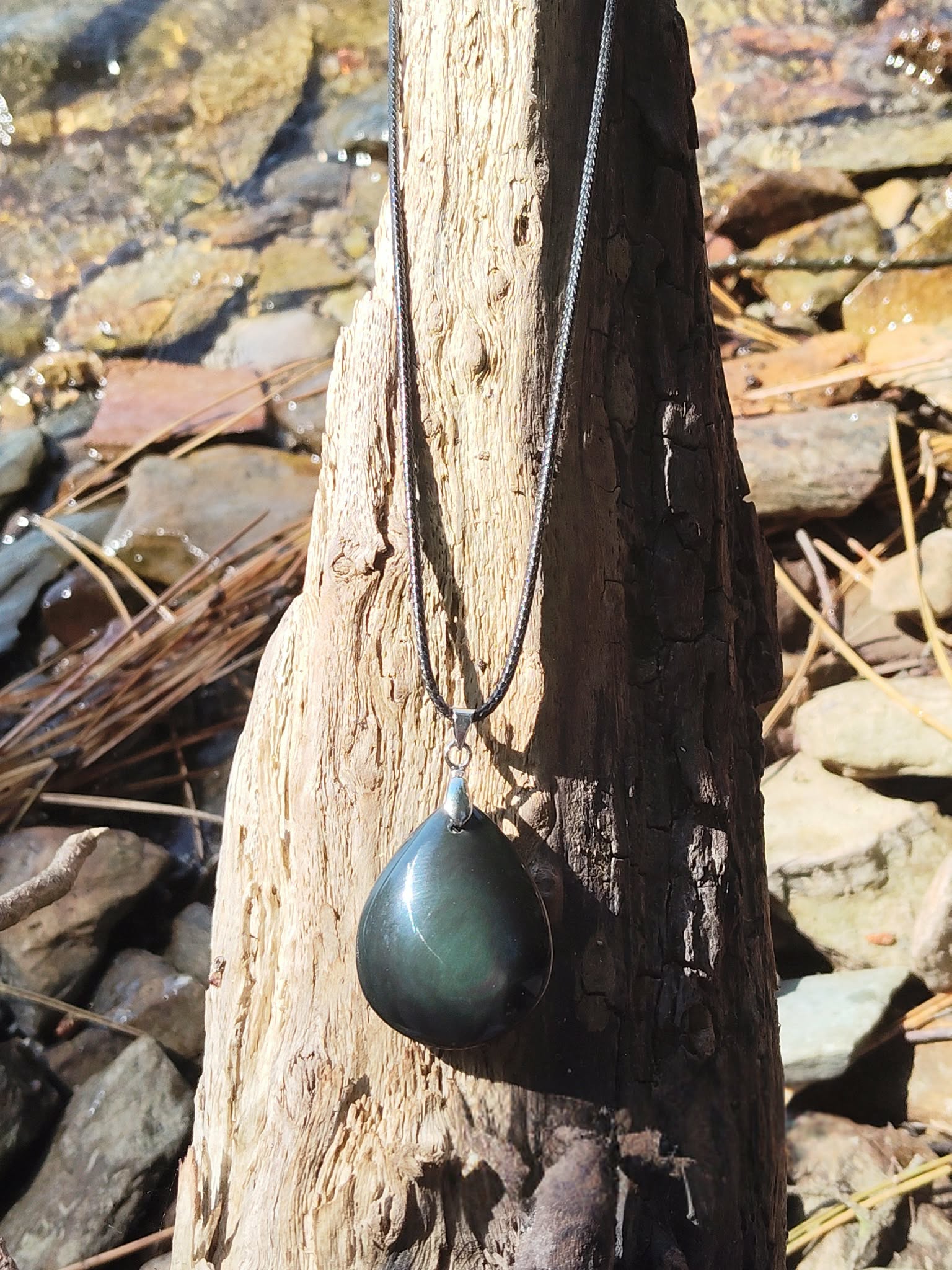 Pendentif obsidienne œil céleste en goutte sur cordon noir, suspendu à un tronc de bois flotté avec galets et eau scintillante en arrière-plan, pierre naturelle associée à l’ancrage, à la présence à soi et à la sérénité.