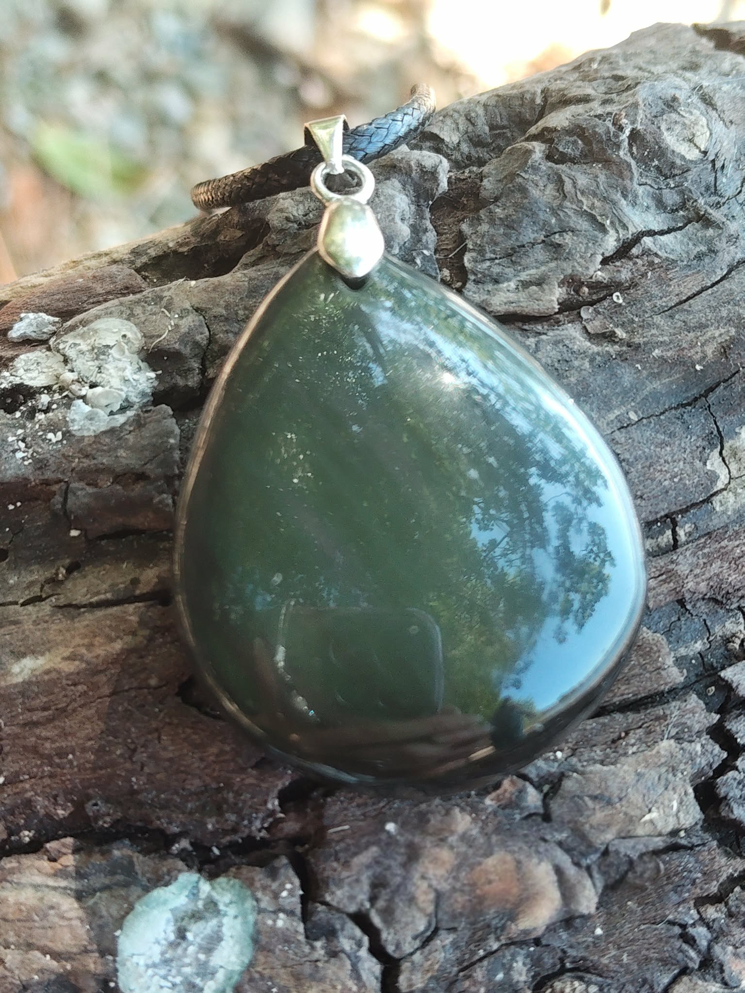 Pendentif goutte en obsidienne œil céleste sur cordon noir, posé sur écorce de bois texturée avec reflets de feuillage, pierre naturelle associée au discernement, au calme profond et à la protection.