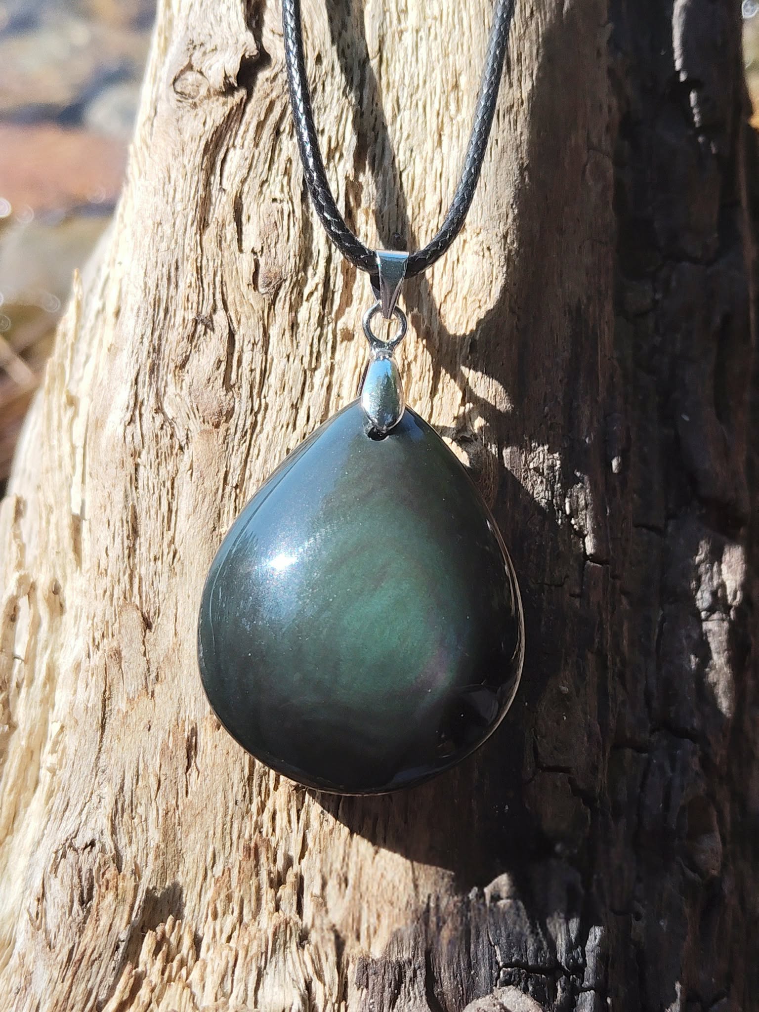 Pendentif obsidienne œil céleste en goutte aux reflets verts sur cordon noir, suspendu à un tronc de bois flotté au bord de l’eau, pierre naturelle liée à la clarté, à l’ancrage et à la protection subtile.