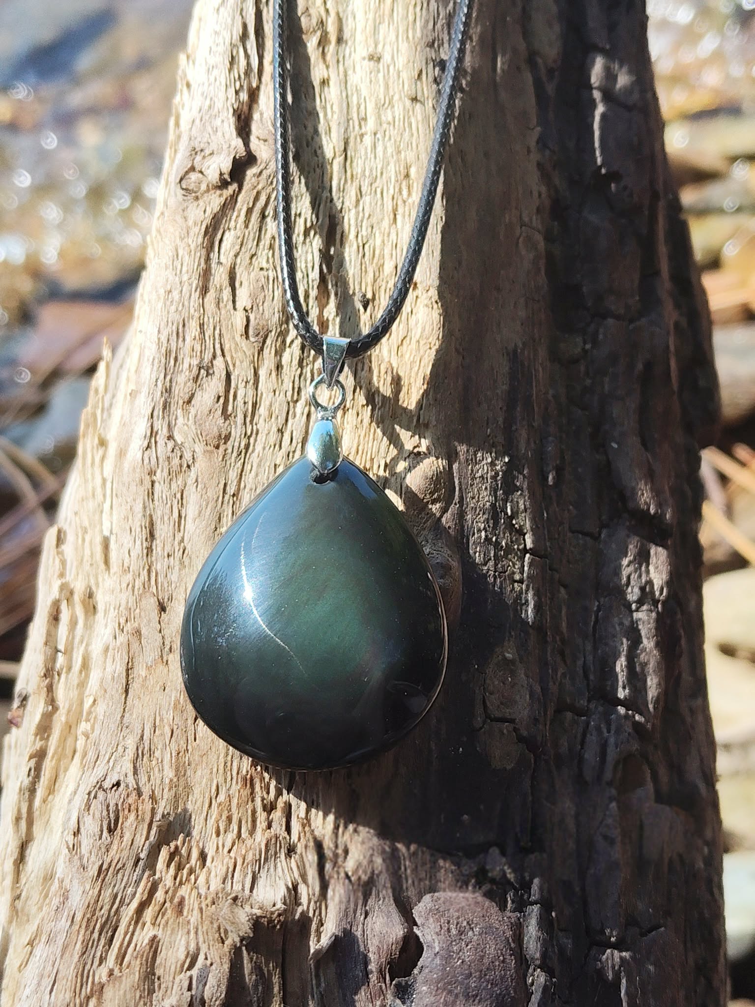 Pendentif obsidienne œil céleste en goutte sur cordon noir, suspendu à un tronc de bois au bord de l’eau, pierre naturelle associée à la protection, à la lucidité et à la stabilité intérieure.