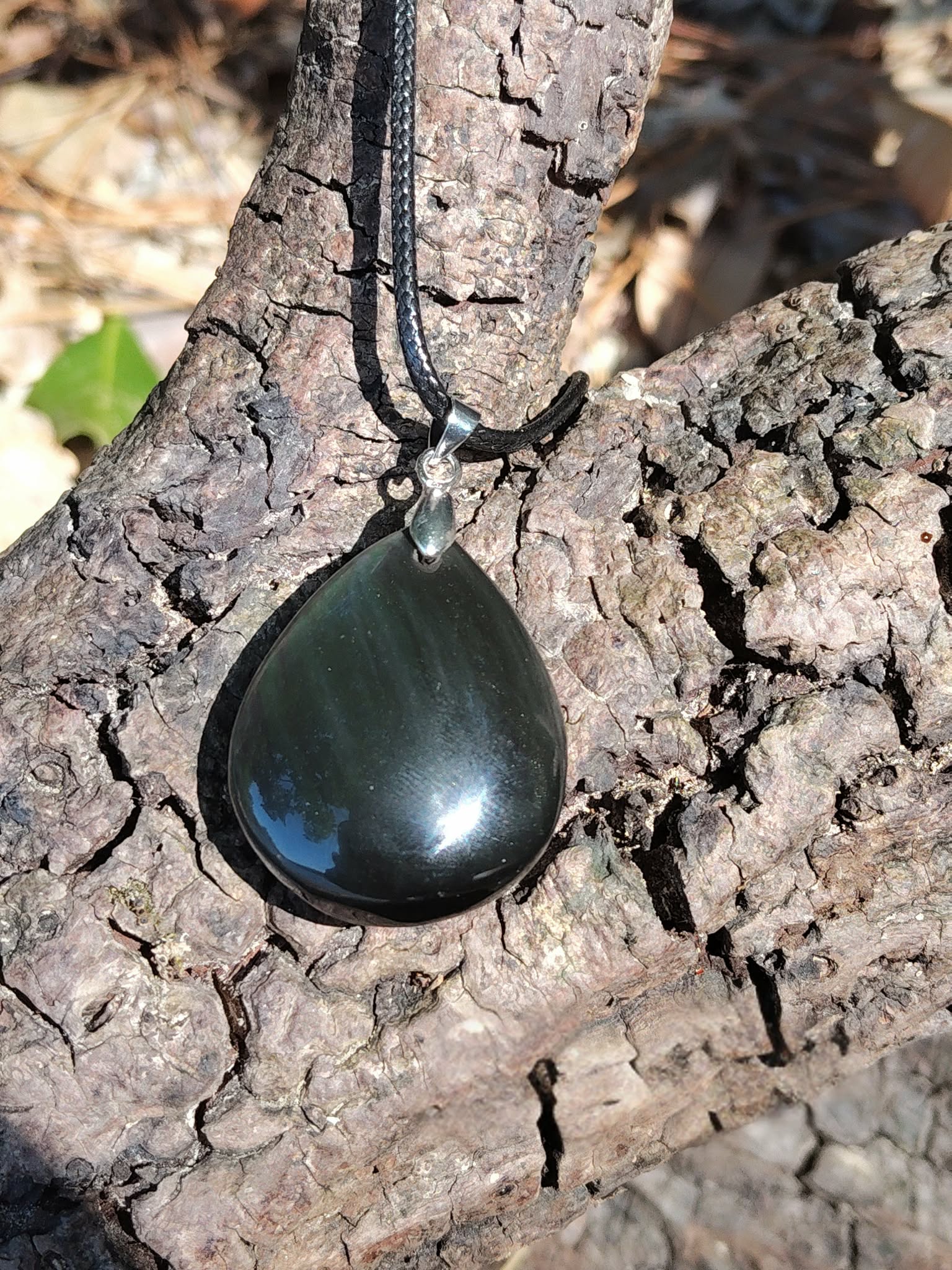 Pendentif obsidienne œil céleste en goutte sur cordon noir, posé sur écorce craquelée au soleil avec reflets profonds, bijou en pierre naturelle évoquant la stabilité, le recentrage et la puissance intérieure.