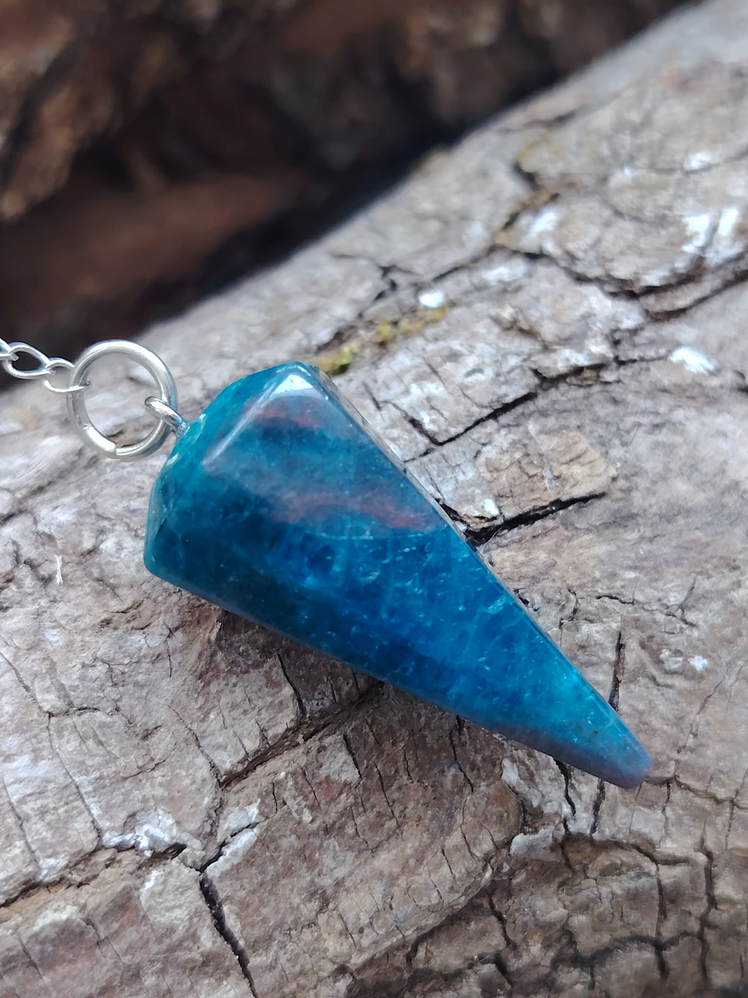 Pendule en apatite bleue facettée posé sur une écorce naturelle aux reliefs marqués, dans une ambiance brute et minérale, évoquant intuition, apaisement émotionnel et clarté intérieure.