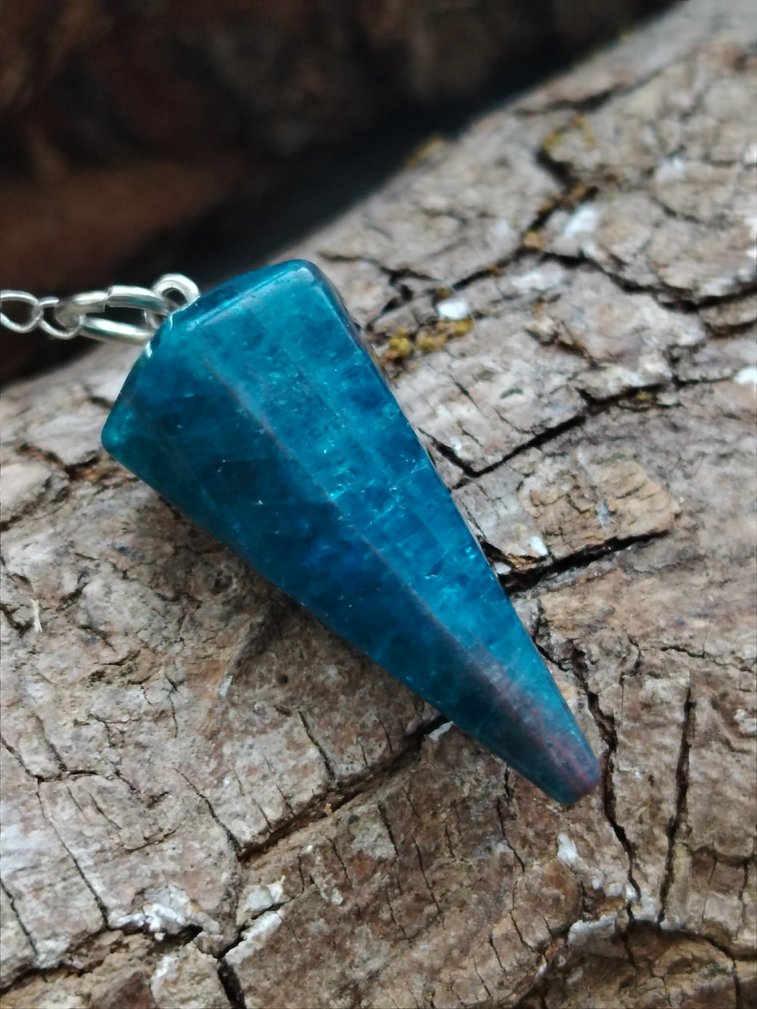 Pendule en apatite bleue facettée posé sur une écorce naturelle aux tons bruns, dans une ambiance minérale et boisée, évoquant intuition, apaisement émotionnel et clarté intérieure.
