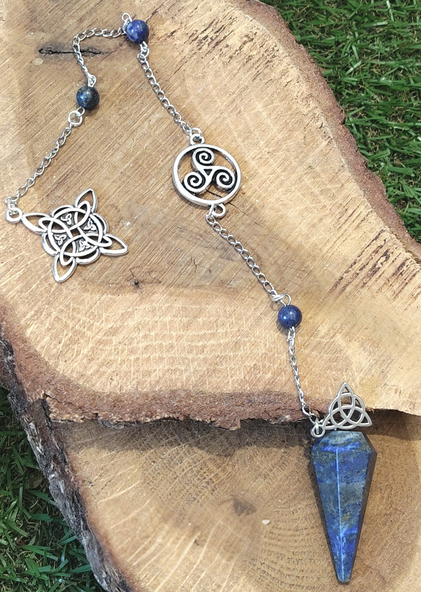Pendule en lapis-lazuli bleu avec chaîne personnalisée argentée et symboles celtiques, présenté sur une tranche de bois dans un décor naturel inspirant recentrage et intuition.