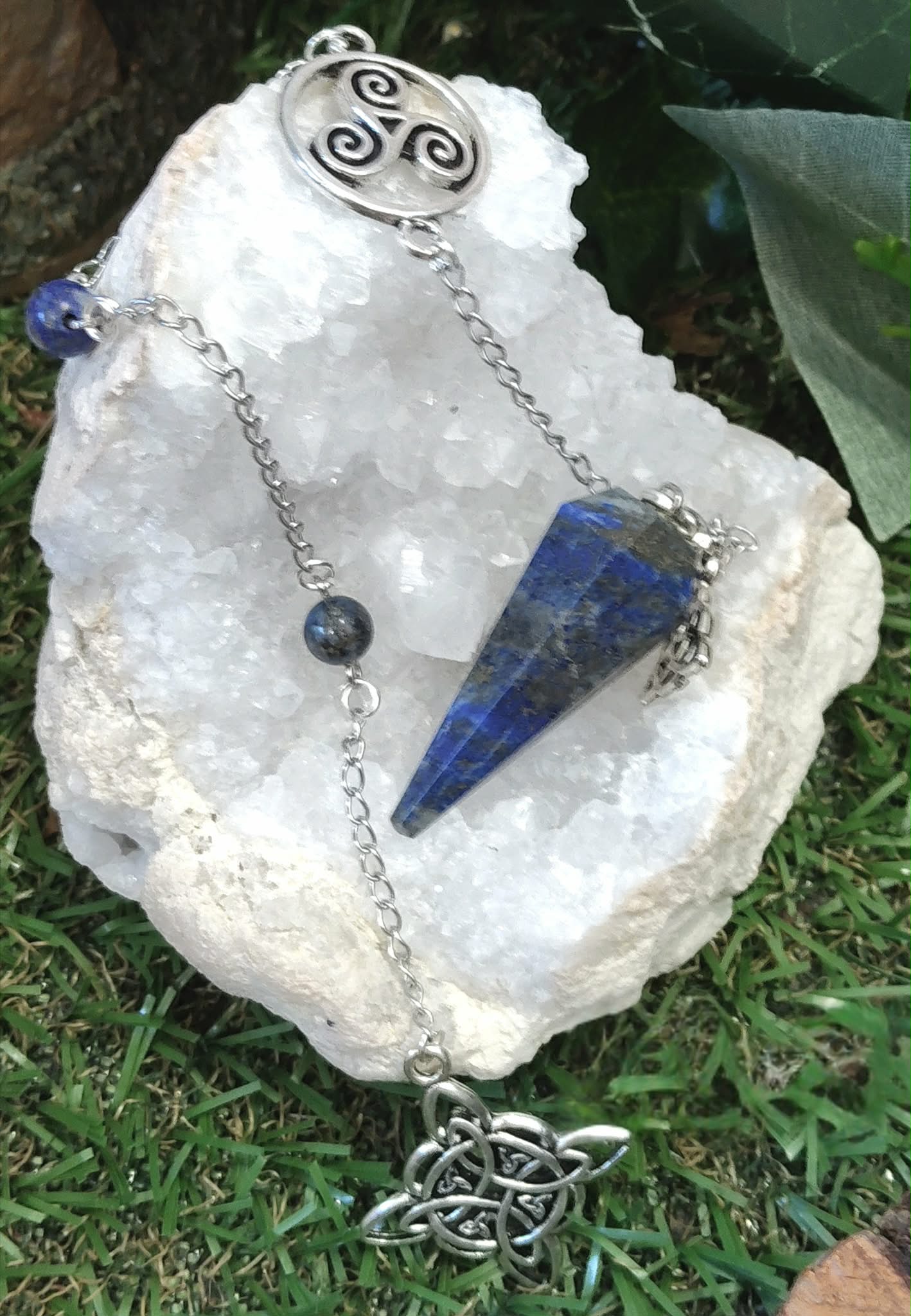 Pendule en lapis-lazuli bleu profond avec chaîne personnalisée par Magic Gaïa, présenté sur une coquille blanche dans un décor naturel évoquant intuition et sérénité.