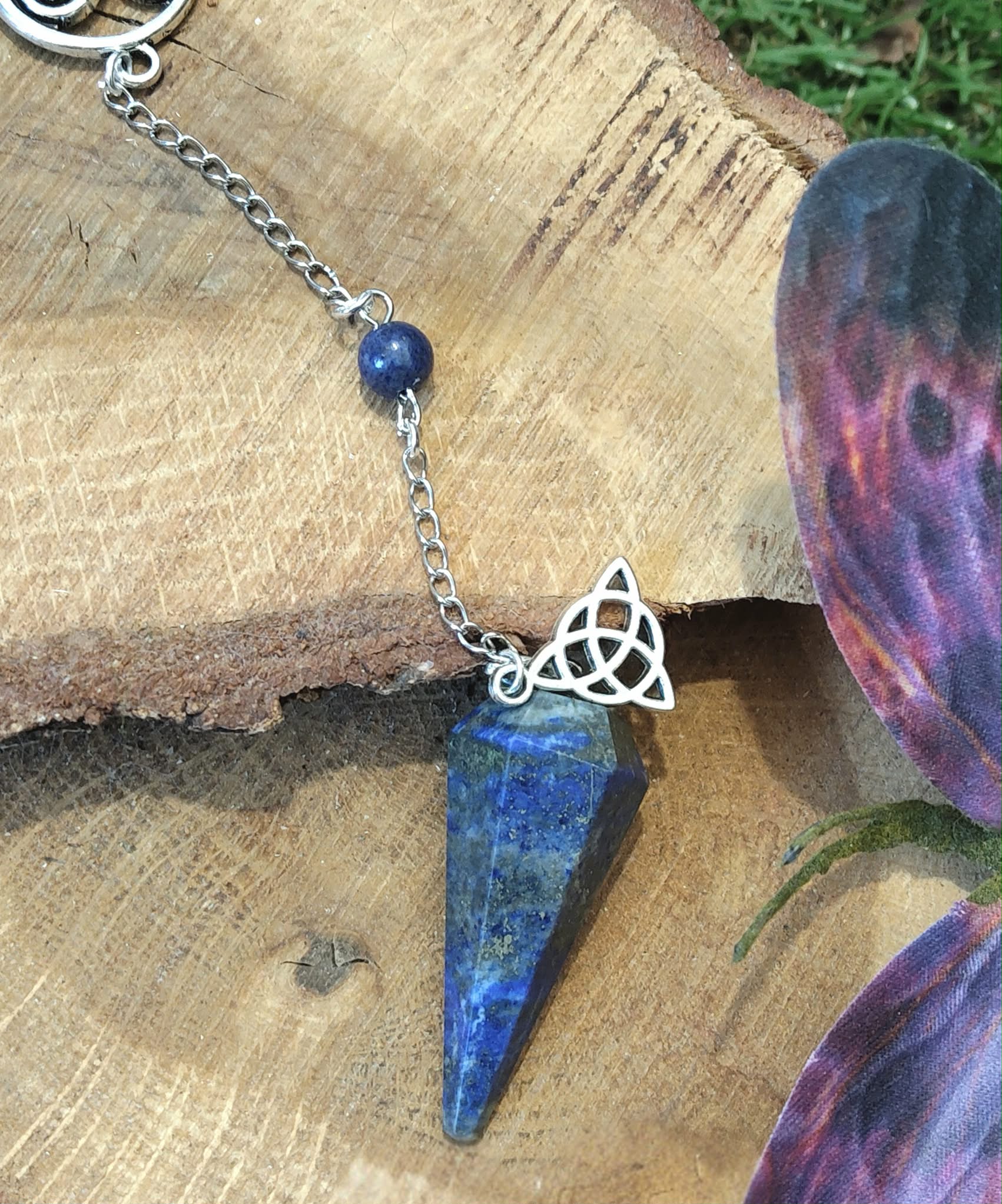 Pendule en lapis-lazuli bleu présenté sur du bois avec chaîne argentée, perle assortie et pierre décorative violette, dans une ambiance naturelle propice à l’intuition.