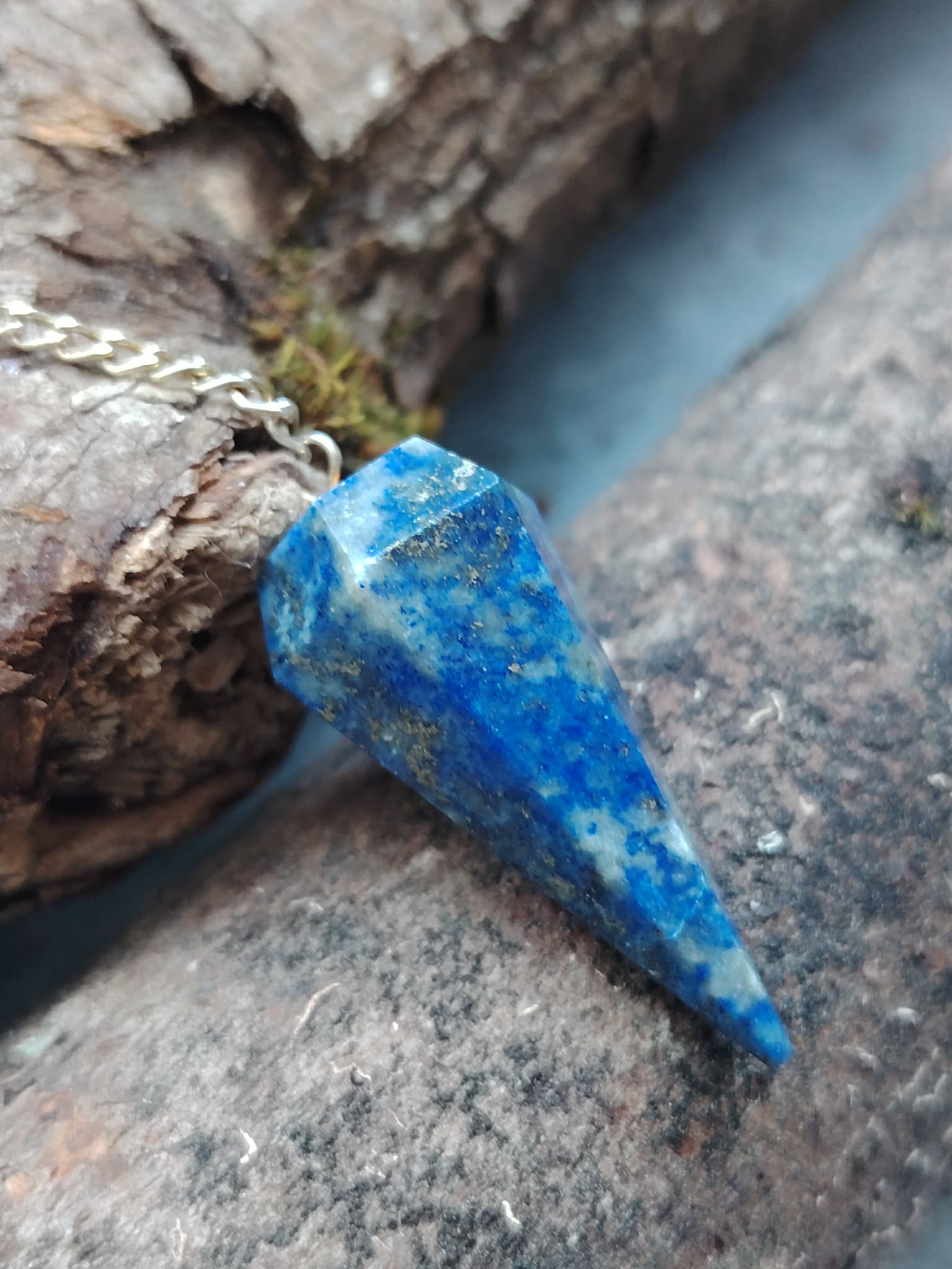 Pendule en lapis-lazuli bleu profond posé entre bois flotté et pierre brute, dans un décor naturel inspirant intuition, clarté mentale et apaisement.