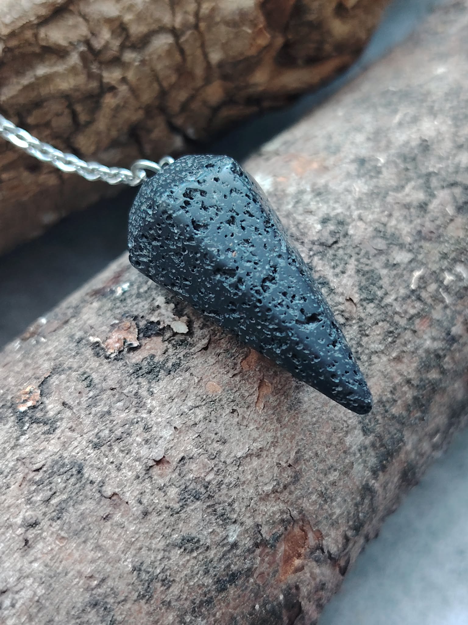 Pendule en pierre de lave noire posé sur une pierre claire, avec bois naturel en arrière-plan, dans un décor brut évoquant ancrage, stabilité et recentrage.