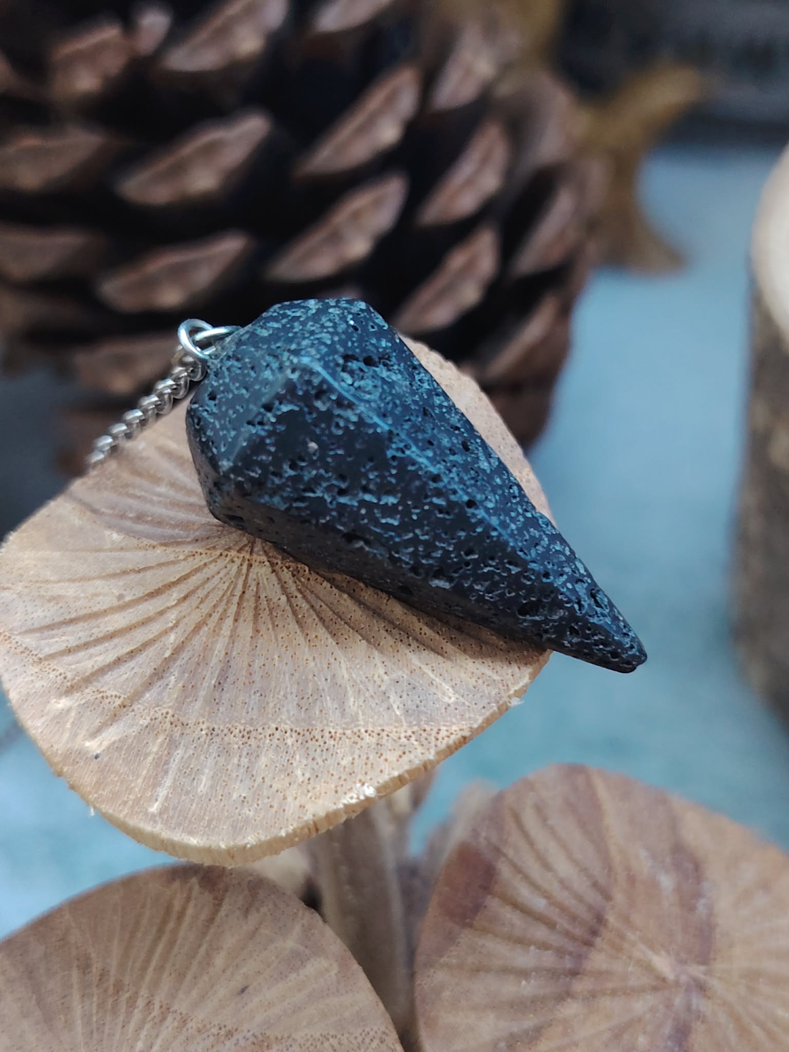 Pendule en pierre de lave noire posé sur un coquillage beige, avec galets et fond bleu dans un décor marin apaisant évoquant ancrage, stabilité et recentrage.