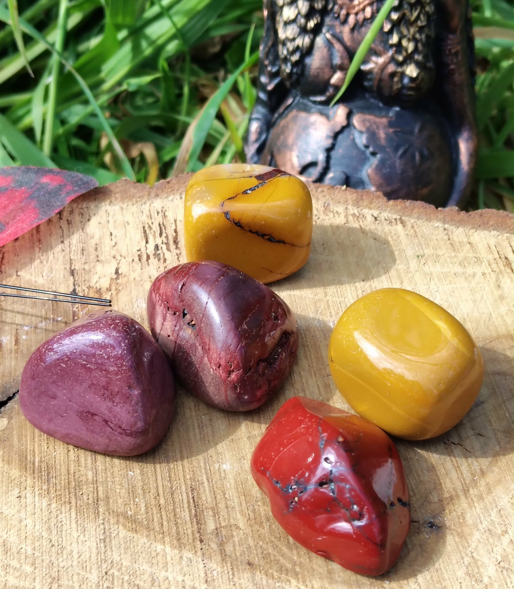 Pierres roulées de jaspe mookaïte posées sur support en bois avec herbes en lumière naturelle, pierres naturelles associées à l’ancrage, à la stabilité intérieure et à l’énergie chaleureuse.