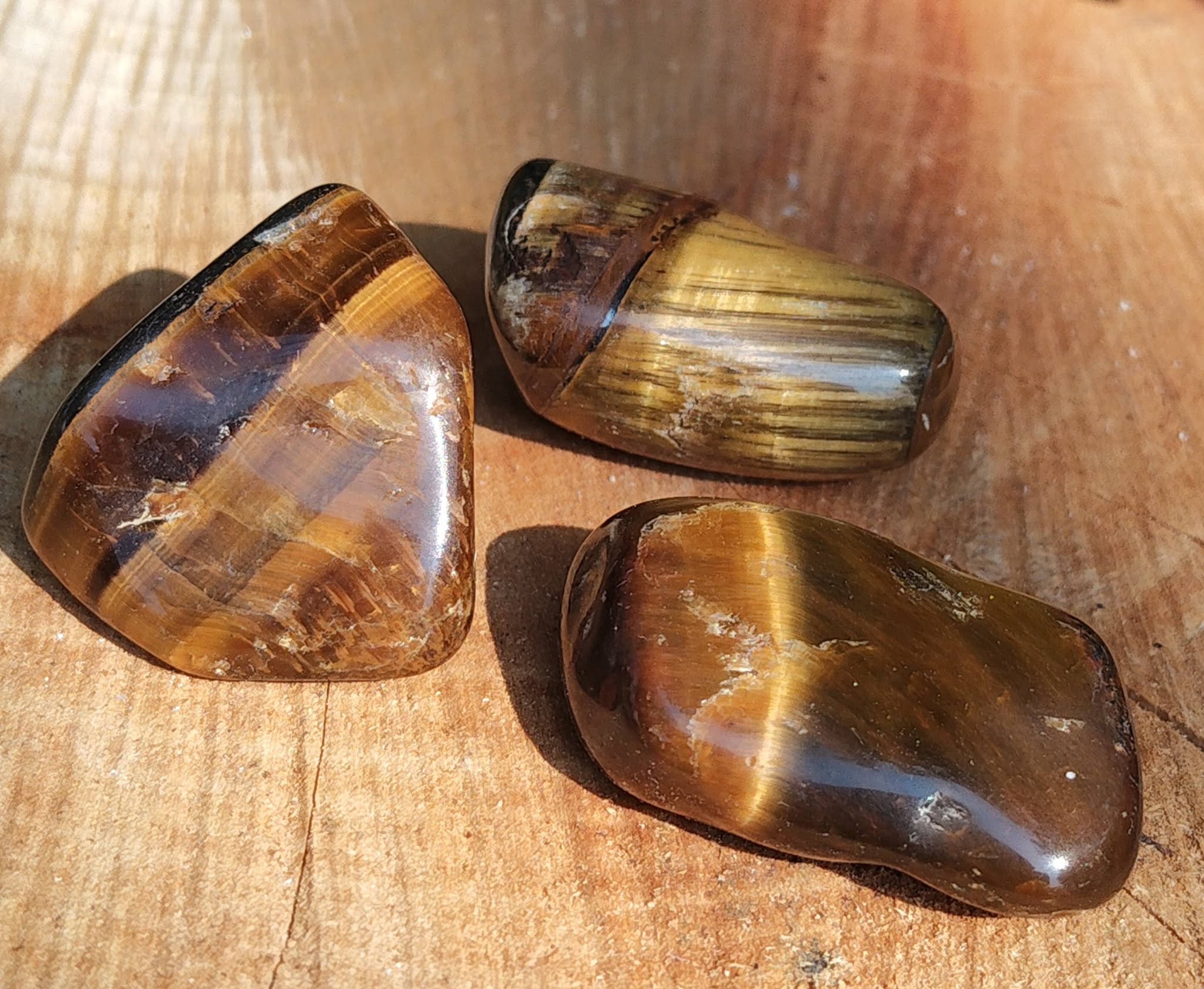 Trois pierres roulées œil de tigre aux reflets dorés et bruns sur fond bois chaleureux, pierres naturelles associées à la stabilité, à l’aplomb et à la présence à soi.