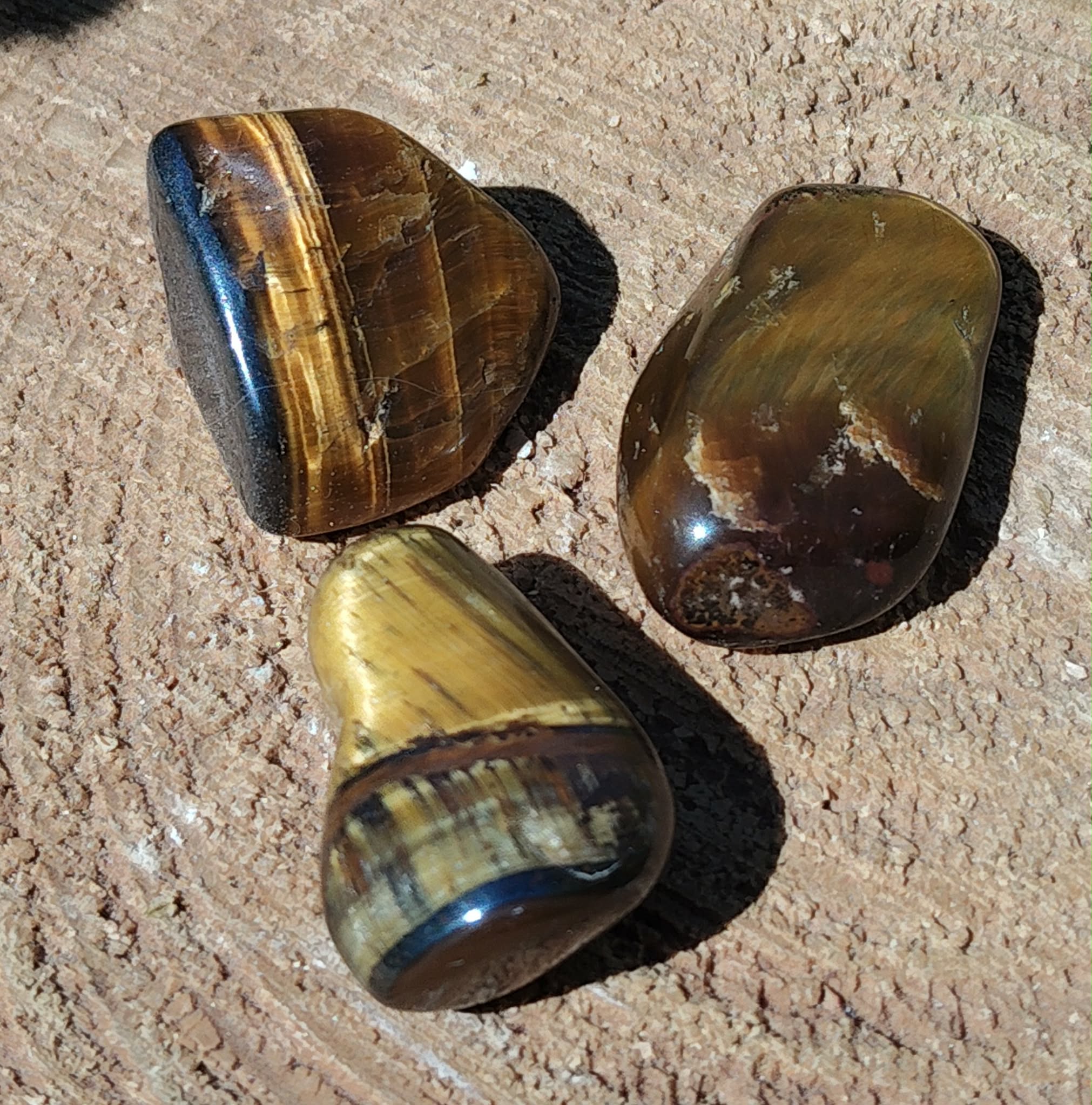 Trois pierres roulées œil de tigre aux reflets bruns et dorés posées sur une surface sableuse, pierres naturelles associées à l’enracinement, à la maîtrise de soi et à l’assurance.
