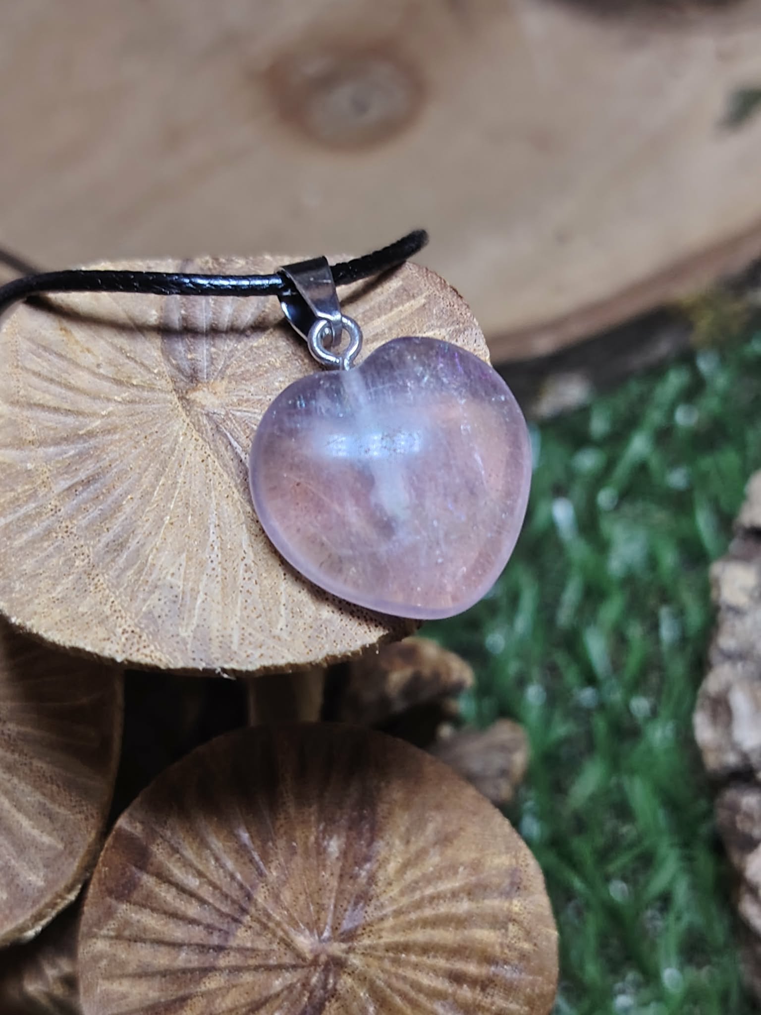 Pendentif cœur en fluorite multicolore aux nuances violettes et bleutées, suspendu sur cordon noir devant un décor végétal, évoquant clarté, apaisement et harmonie intérieure.