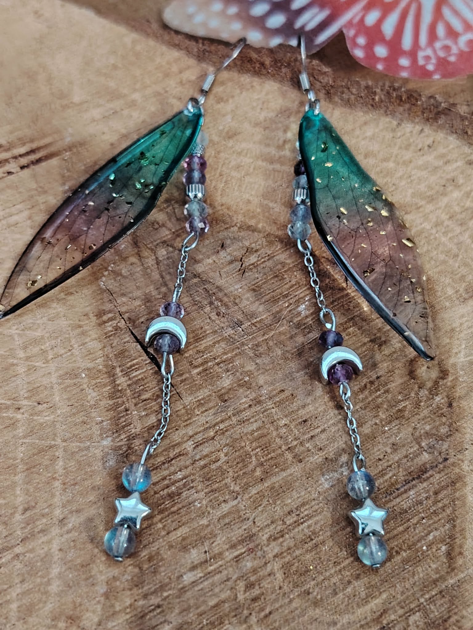 Boucles d’oreilles fée création Magic Gaïa posées sur bois brut, avec ailes translucides bleu-vert, pampilles argentées, lunes et étoiles, dans une ambiance féerique, douce et inspirante au quotidien.
