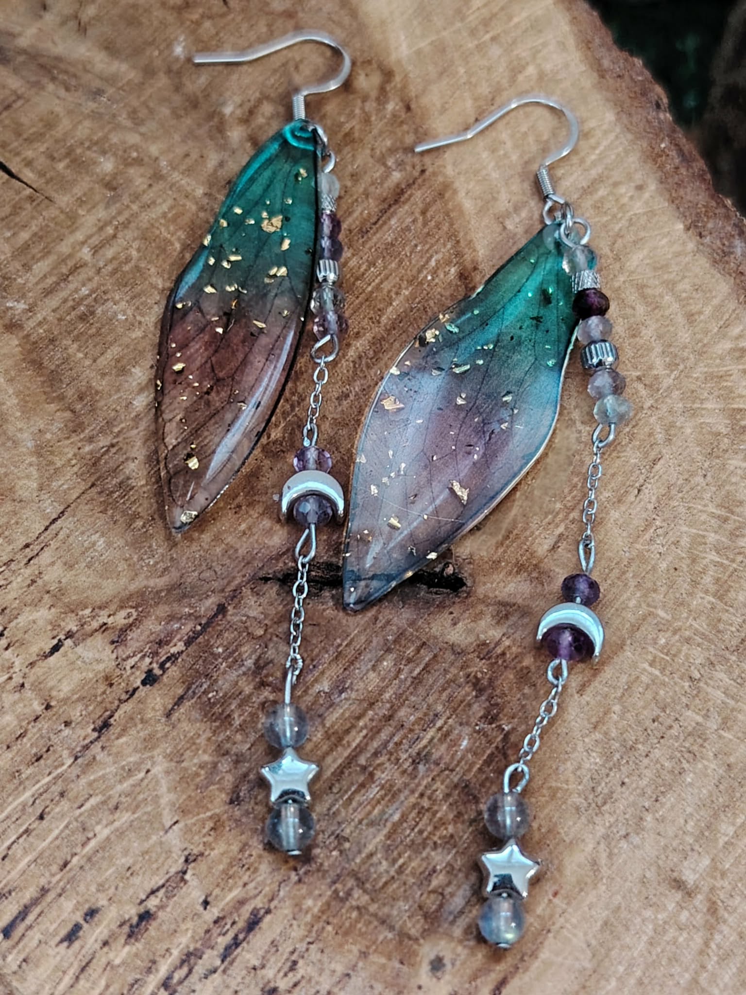Boucles d’oreilles fée création Magic Gaïa posées sur bois brut, avec ailes translucides bleu-vert, pampilles argentées, lunes et étoiles, dans une ambiance féerique, douce et pleine de poésie.