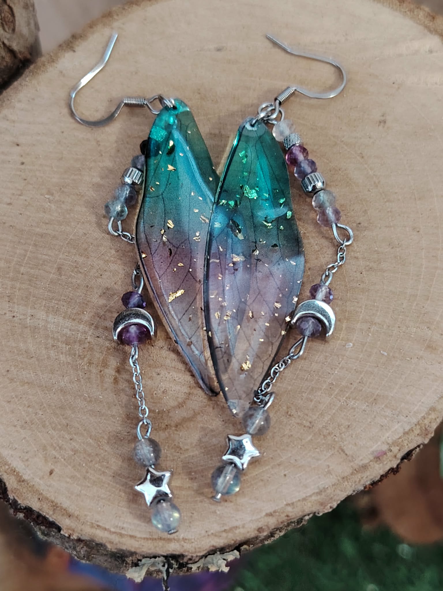 Boucles d’oreilles fée création Magic Gaïa posées sur une tranche de bois, avec ailes translucides bleu-vert, fines pampilles argentées, lunes et étoiles, dans une ambiance féerique inspirant douceur et légèreté.