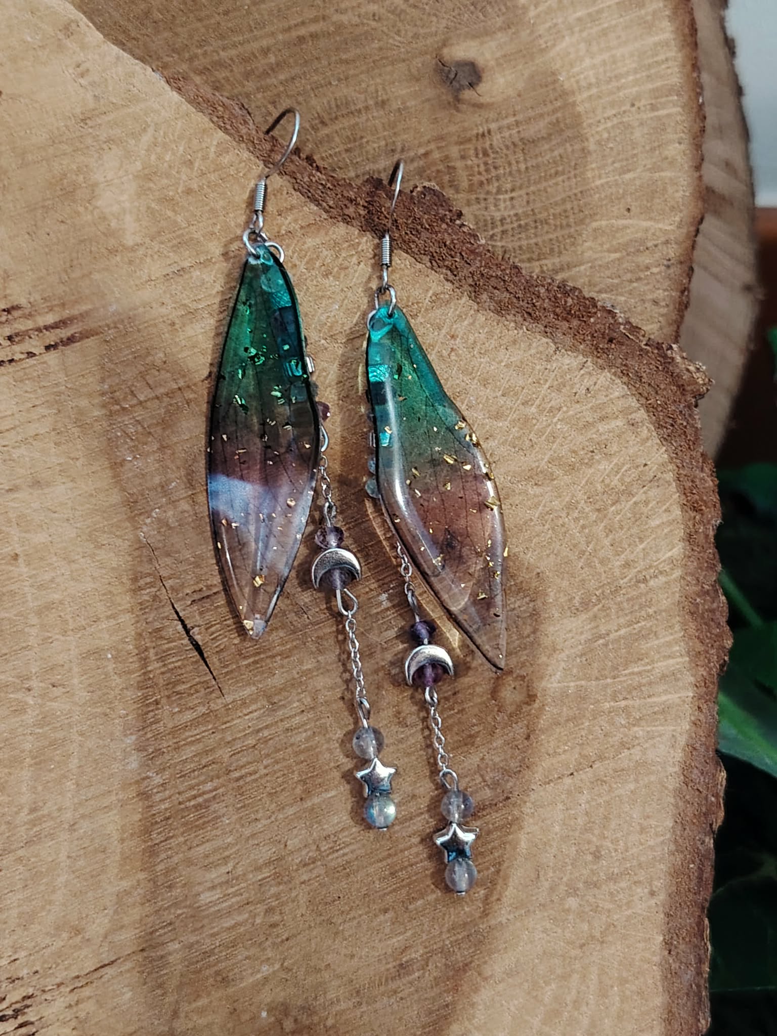 Boucles d’oreilles fée création Magic Gaïa posées sur bois naturel, avec ailes translucides bleu-vert, pampilles lunes et étoiles, dans une ambiance féerique et apaisante propice à la douceur.