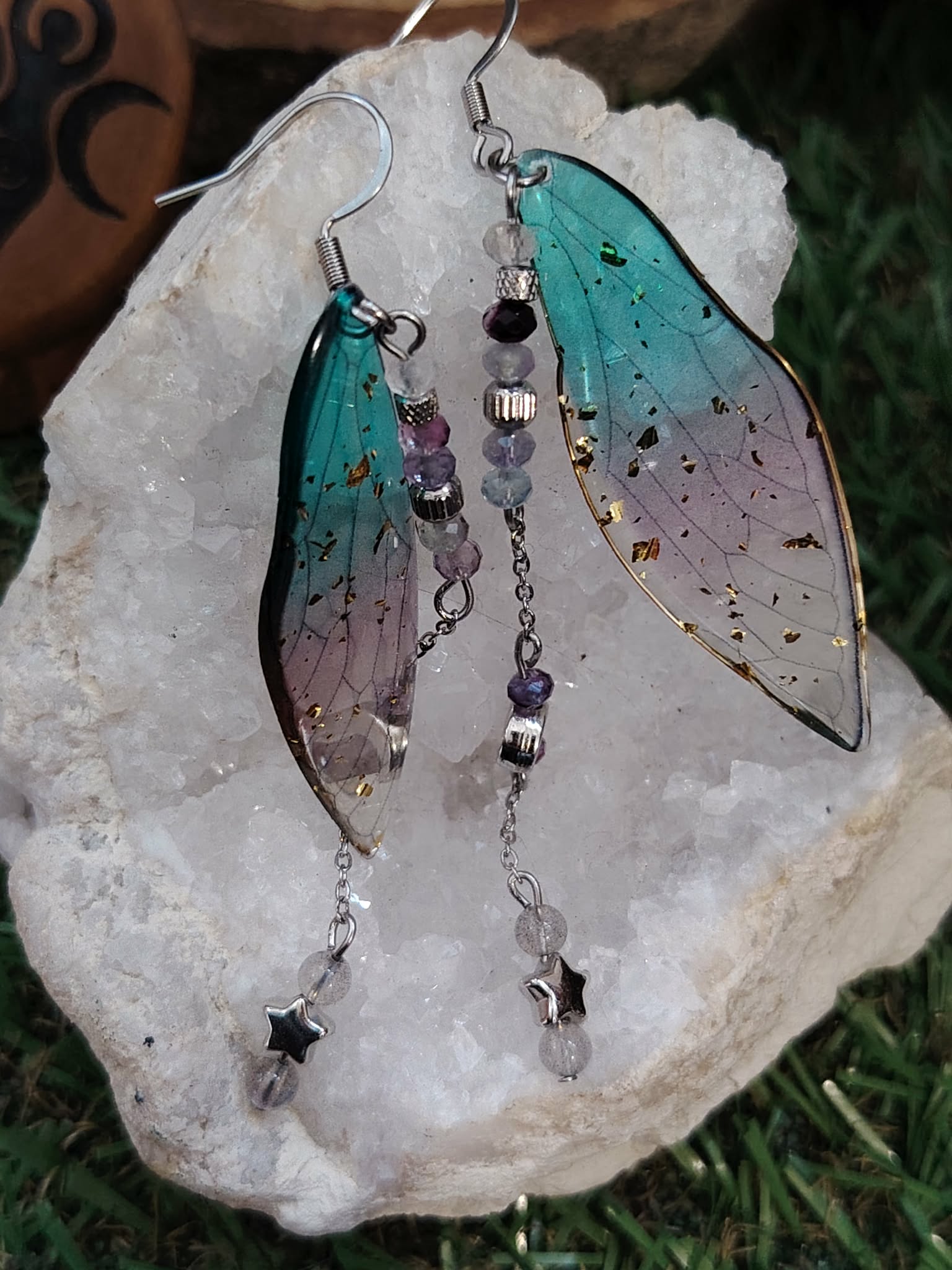 Boucles d’oreilles fée création Magic Gaïa posées sur un amas de quartz, avec ailes translucides bleu-vert, chaînettes fines et perles sombres, pour une ambiance poétique, apaisante et féerique