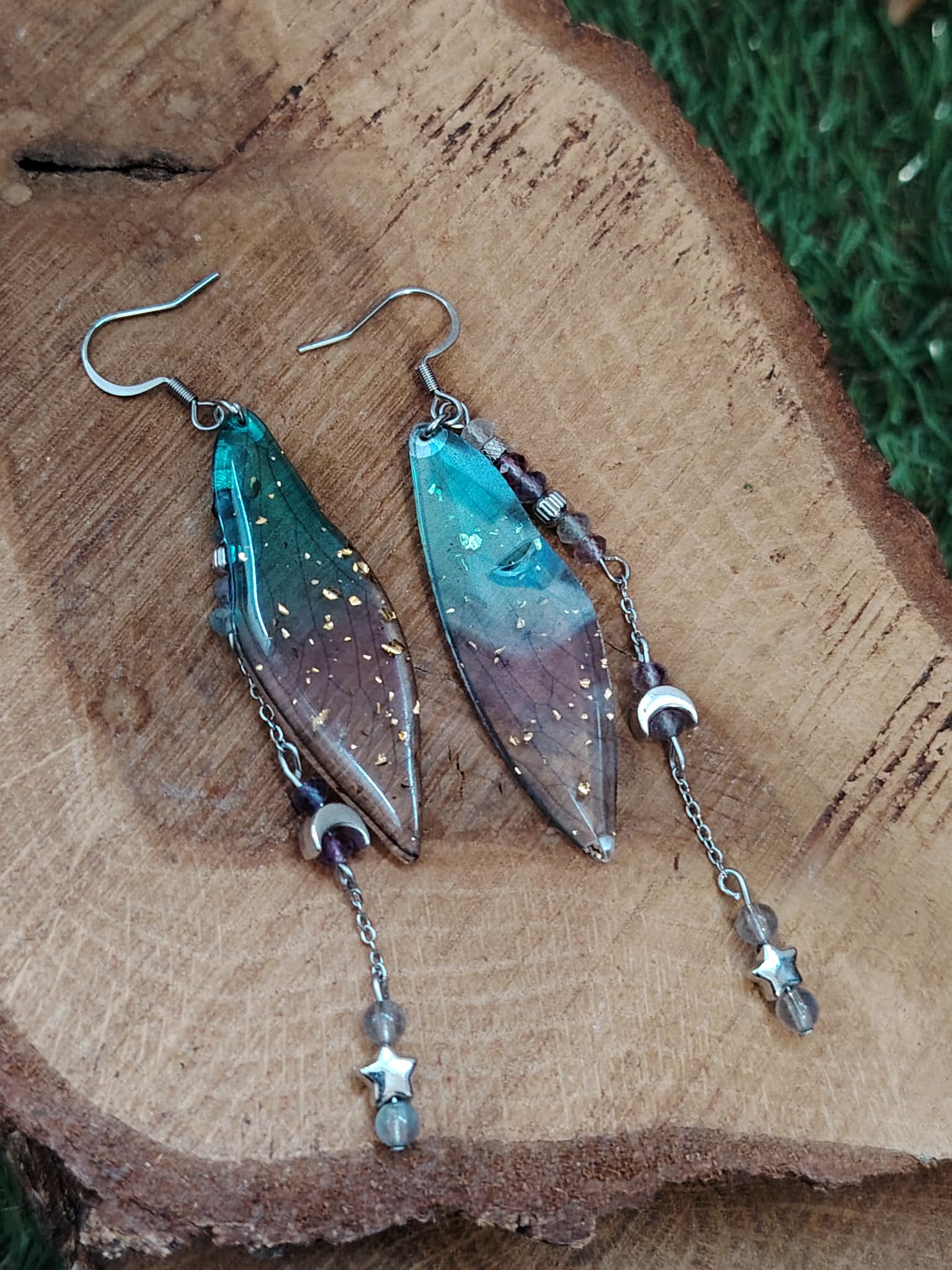 Boucles d’oreilles fée création Magic Gaïa posées sur bois naturel, avec ailes translucides bleu-vert et pampilles fines, dans une ambiance féerique, douce et inspirante favorisant l’évasion et la légèreté.