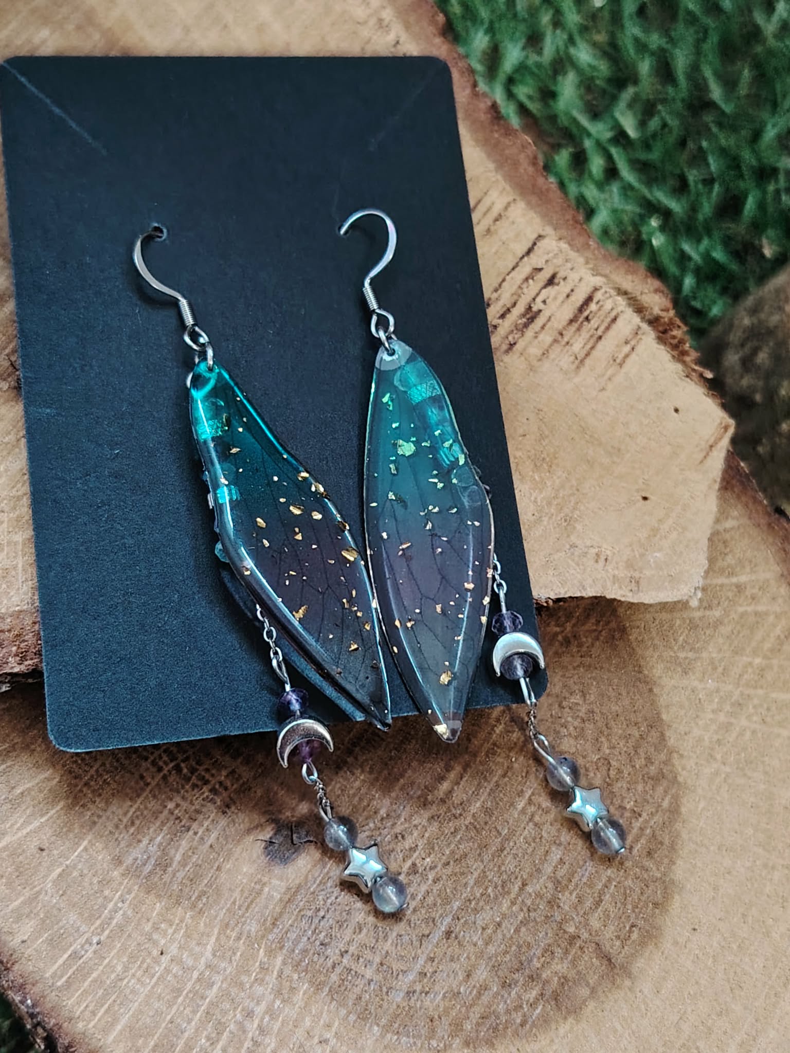 Boucles d’oreilles fée création Magic Gaïa présentées sur support noir, posées sur bois avec mousse verte, ailes bleu-vert irisées et pampilles fines, dans une ambiance féerique évoquant douceur et légèreté.