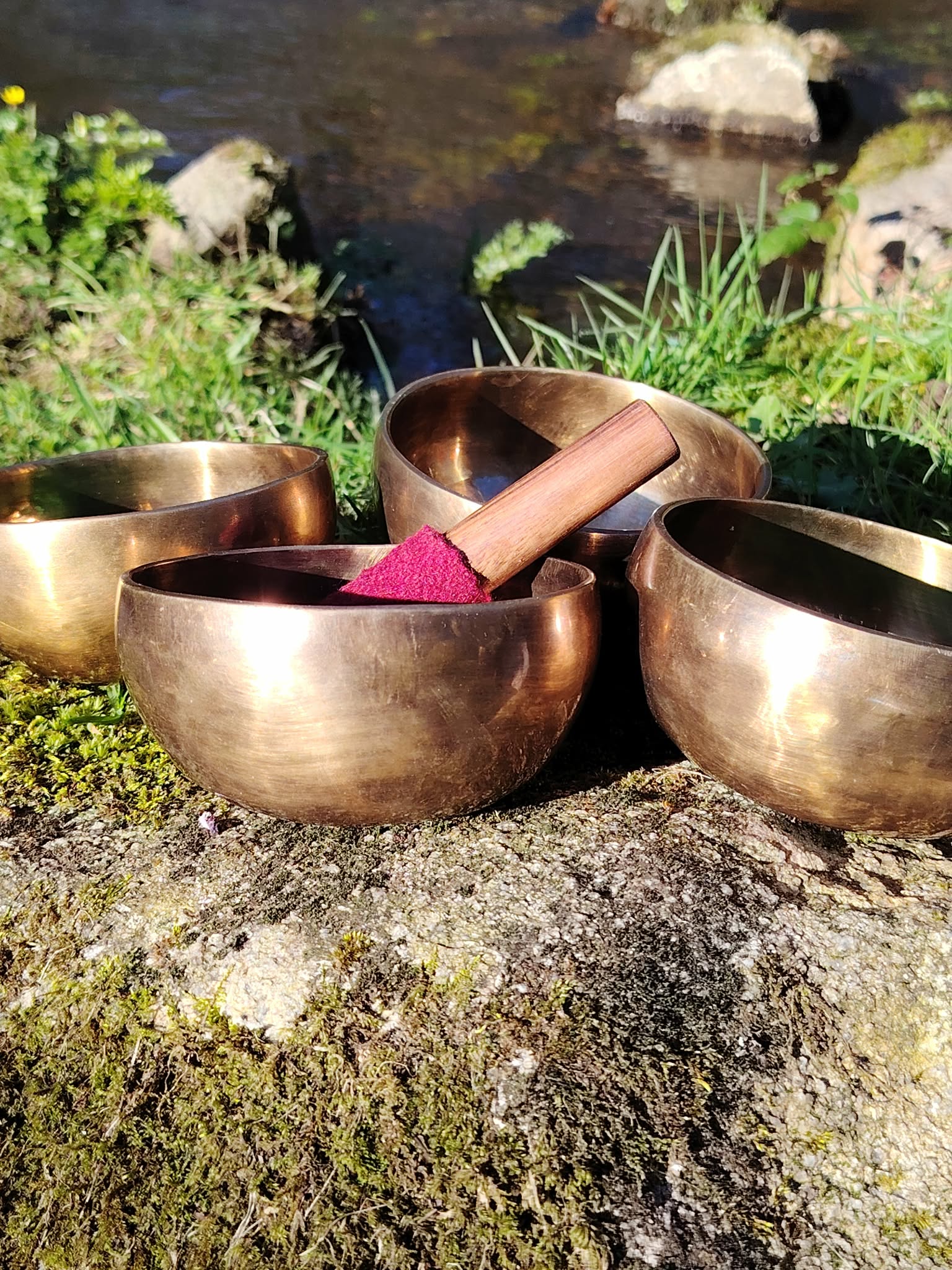 En Bretagne, trio de bols tibétains dorés sur rocher moussu au bord d’un ruisseau, maillet en bois au feutre rouge, pour apaiser et recentrer en douceur.