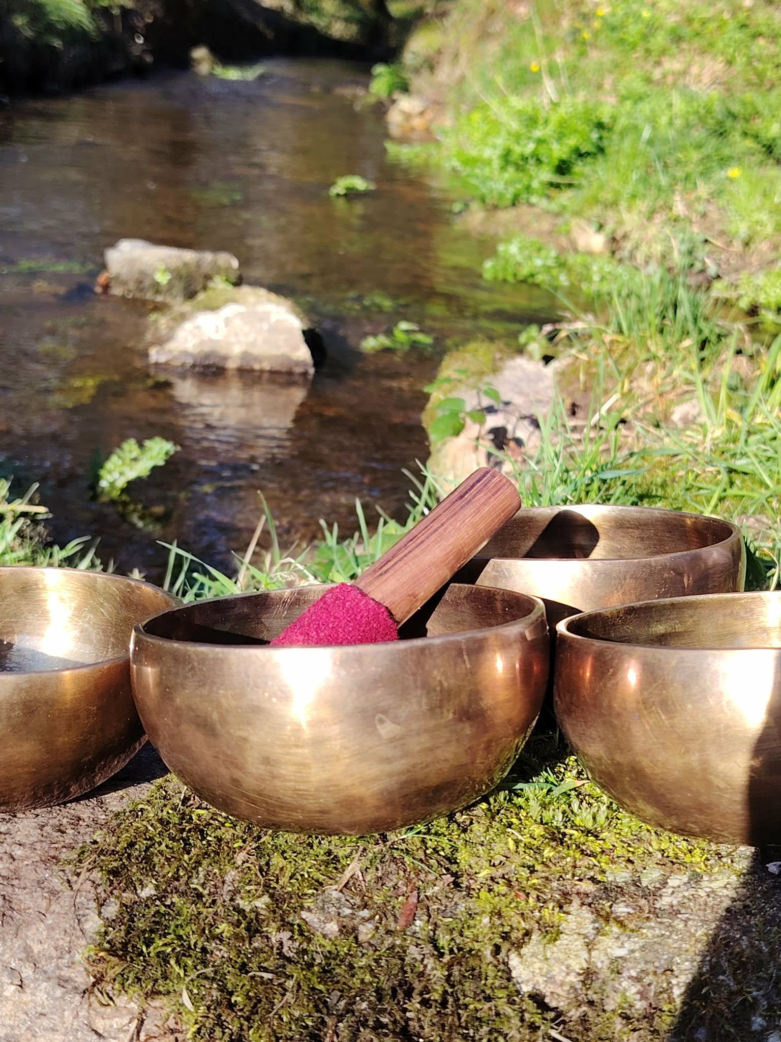 Bols tibétains dorés posés sur un rocher moussu au bord d’un ruisseau, maillet en bois et feutre rouge, ambiance nature pour apaiser l’esprit, recentrer et harmoniser en douceur.