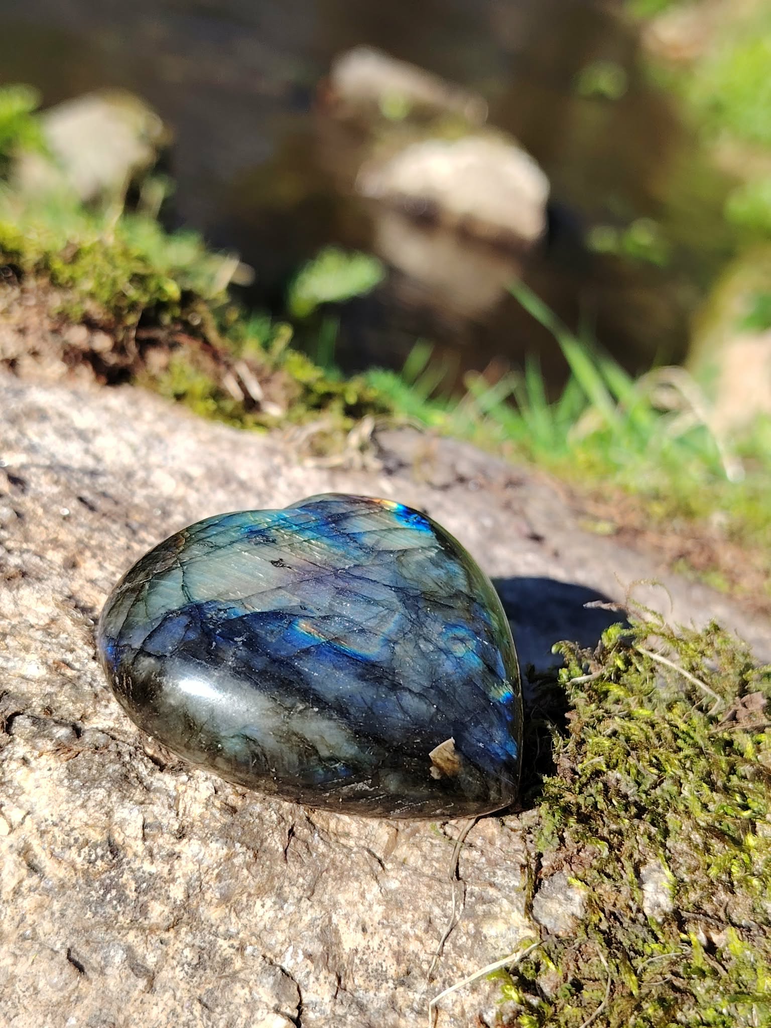 Coeur labradorite poli aux reflets bleus posé sur granit breton avec mousse et ruisseau en arrière-plan, pour protéger l’aura, apaiser et soutenir l’intuition.