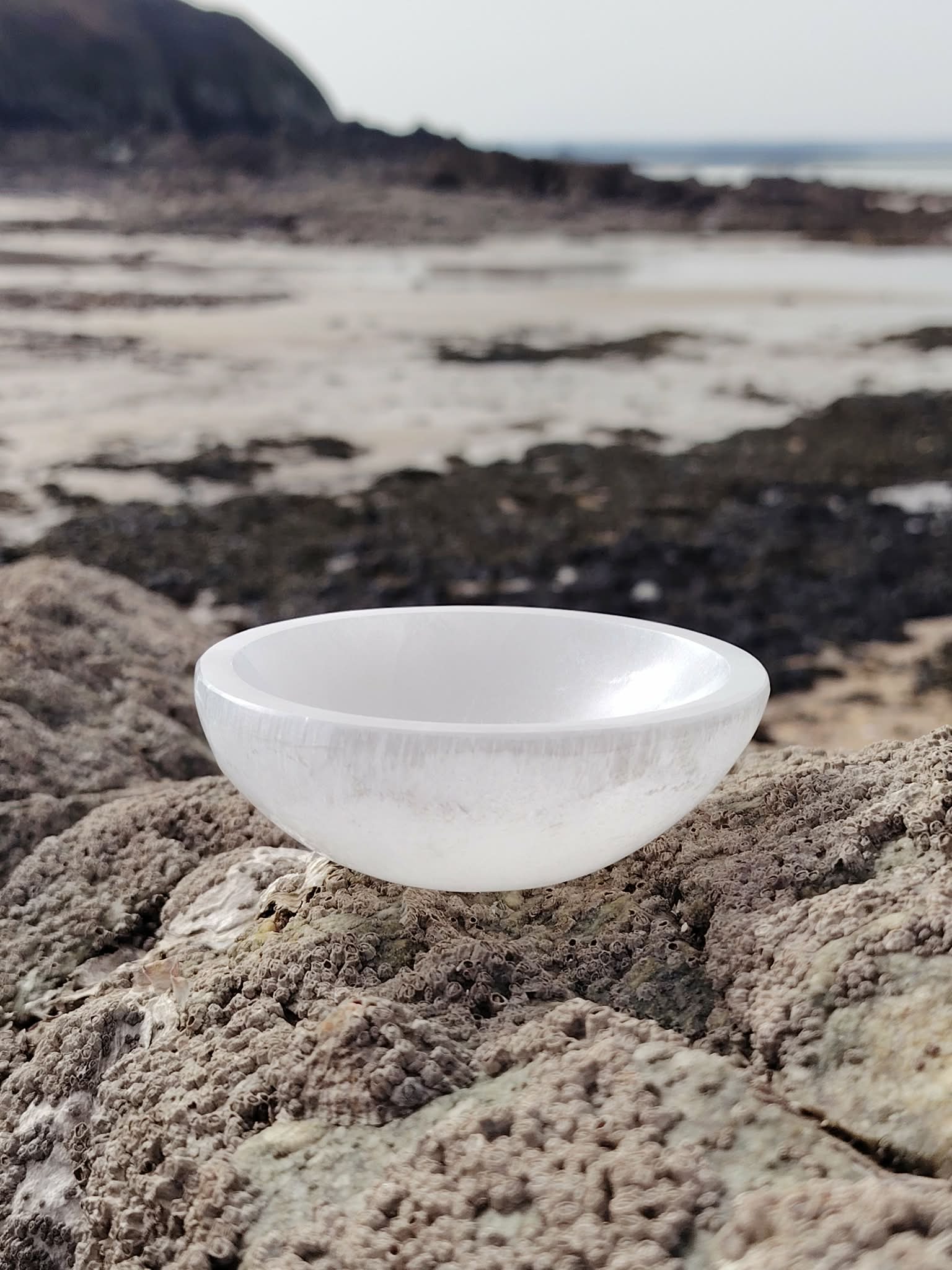 Bol chantant en sélénite 10 cm posé sur rocher granitique face à la mer en Bretagne, vagues et îlot au loin, pour une atmosphère sereine favorisant apaisement et clarté.