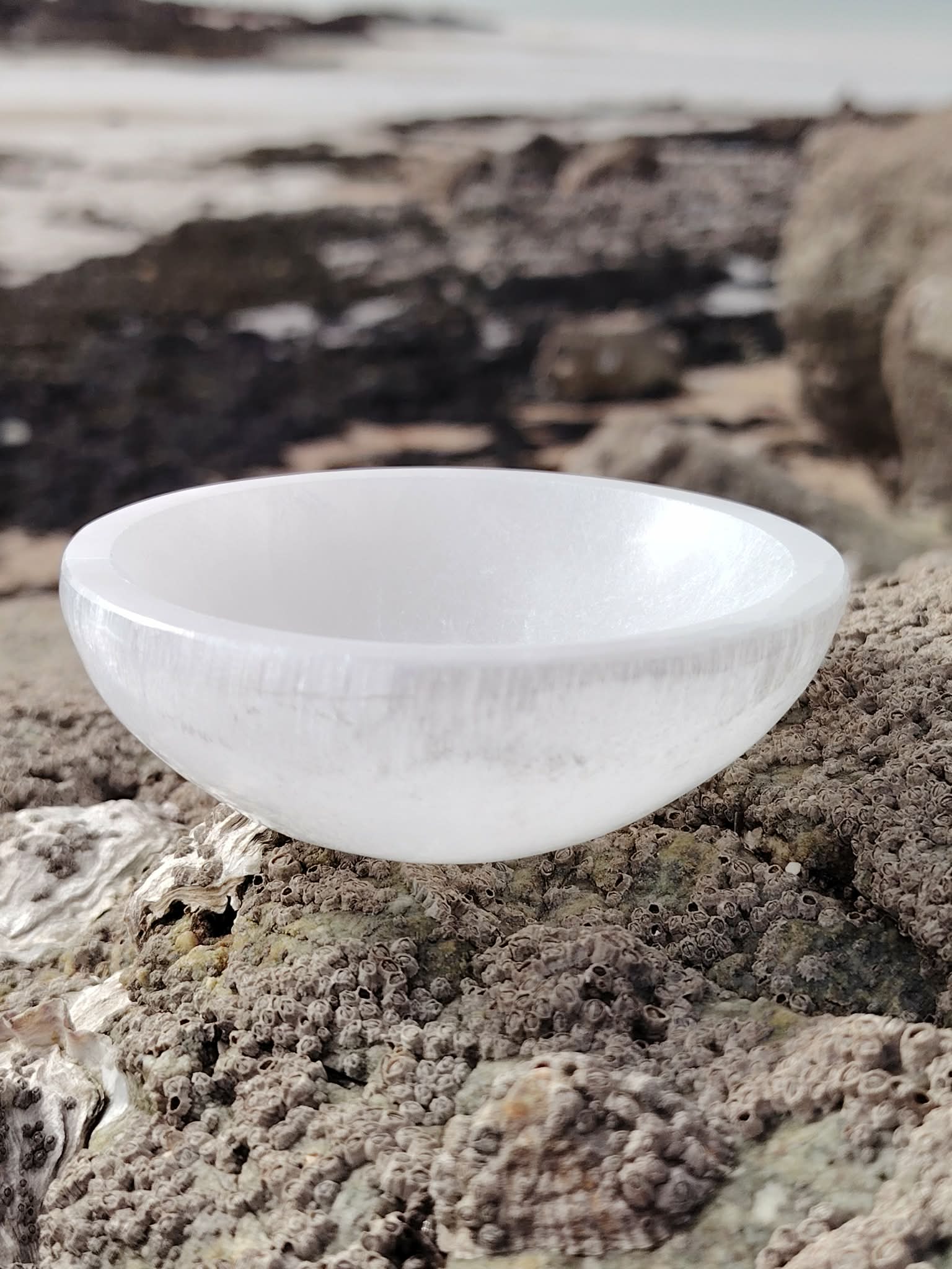 Bol chantant en sélénite 10 cm posé sur un rocher granitique face à la mer en Bretagne, galets et ciel voilé en décor, pour une ambiance apaisante propice à la clarté intérieure et au recentrage.