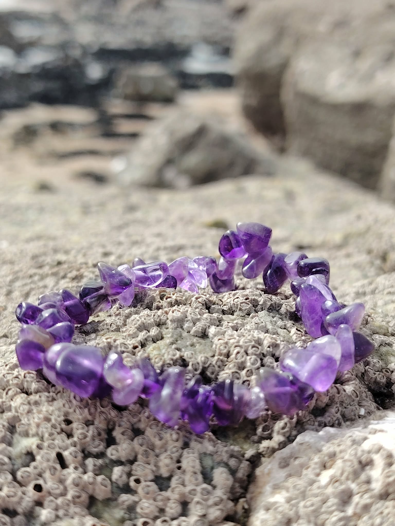 Bracelet améthyste violette sur rocher de granit avec galets et mer floutée, pierre naturelle associée au recentrage et à l’intuition
