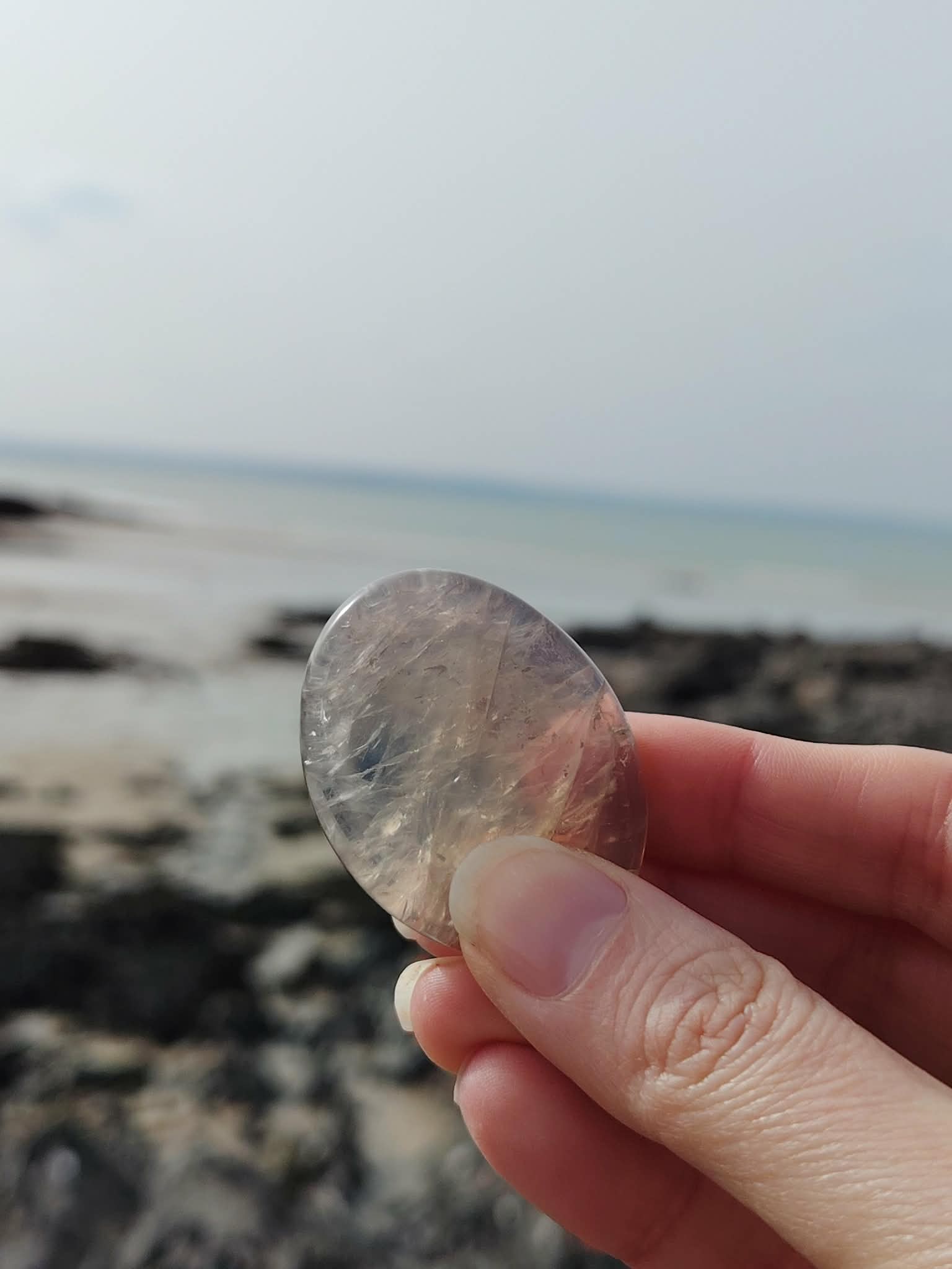 Galet en quartz rose bleu naturel tenu à la main face à la mer en Bretagne, pierre naturelle évoquant le réconfort, l’écoute de soi et la tranquillité émotionnelle.