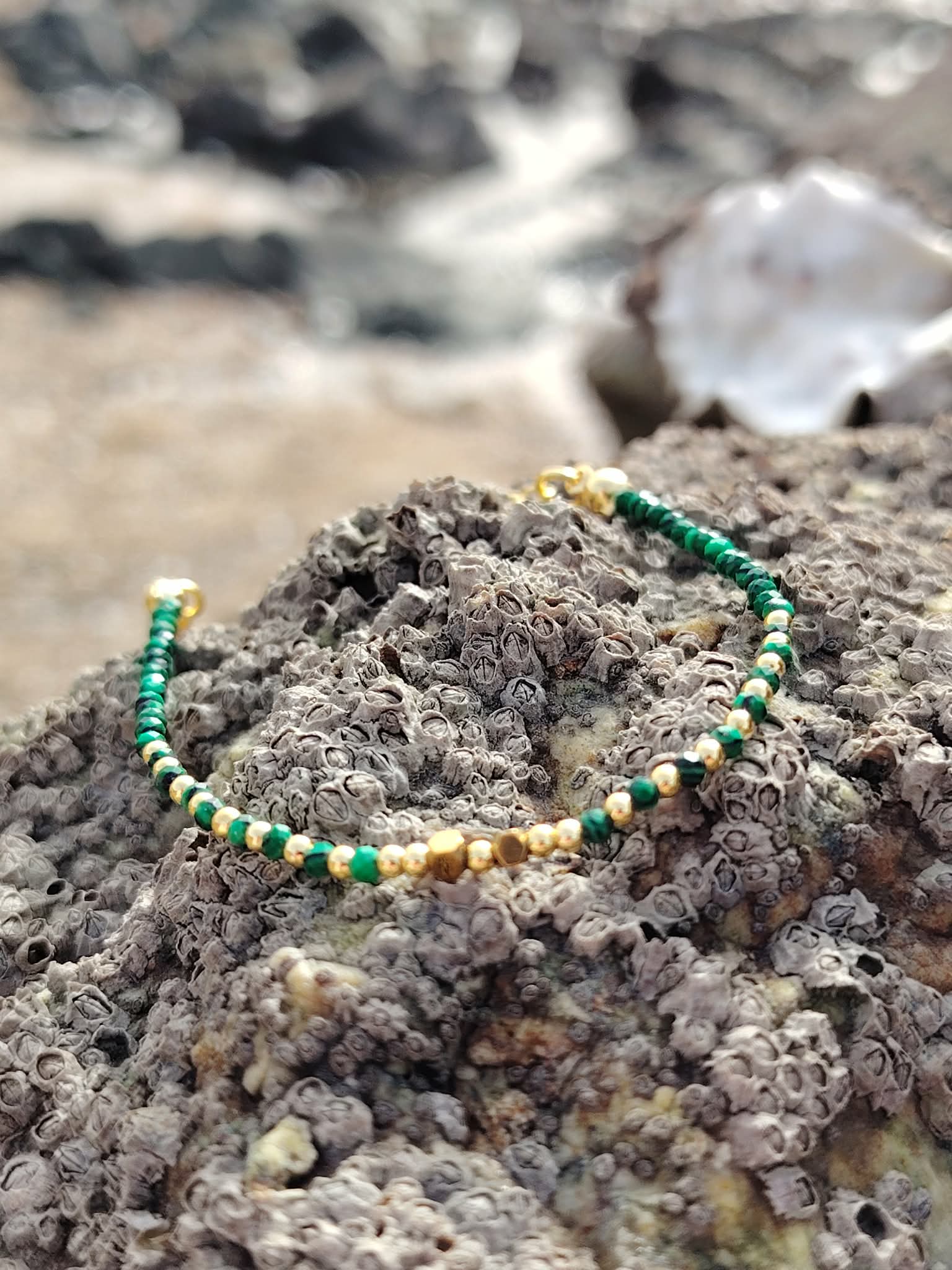 Bracelet malachite facettée 2 mm et cuivre plaqué or sur rocher en Bretagne, plage et coquillage en arrière-plan, bijou vert lumineux