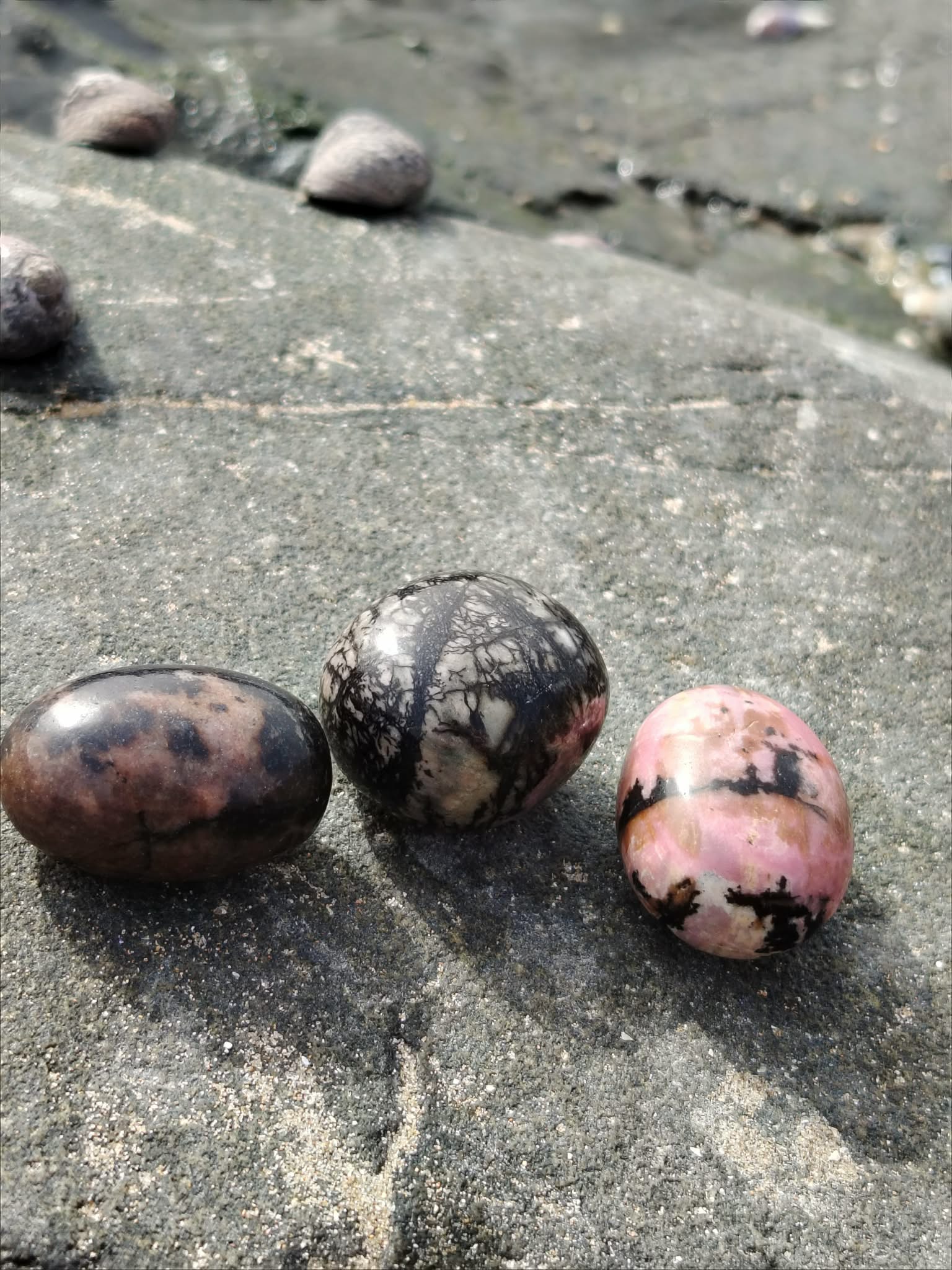 Trois pierres roulées rhodonite aux teintes rose et noir posées sur la roche en Bretagne, évoquant apaisement, ancrage et équilibre émotionnel.