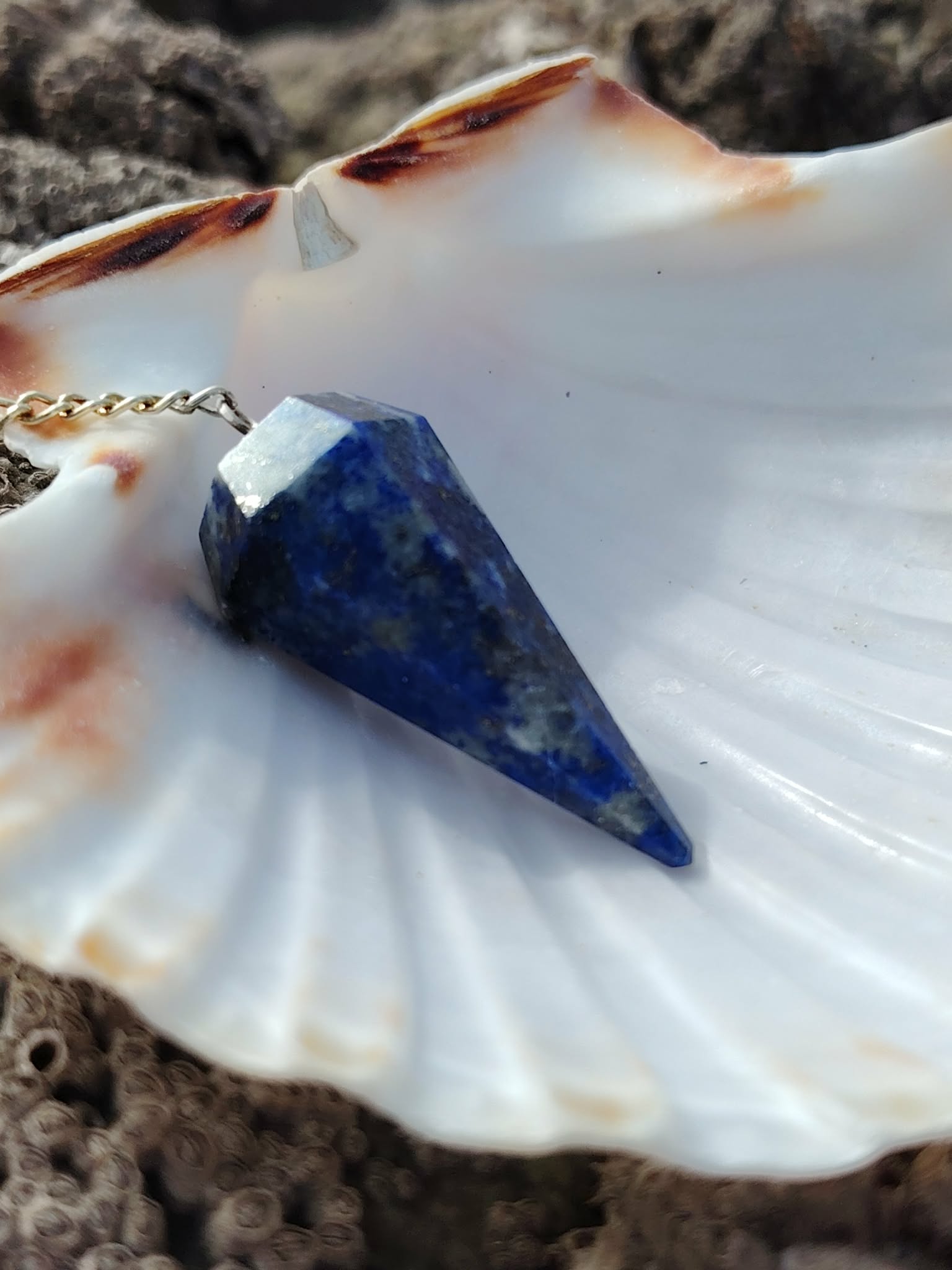 Pendule en lapis-lazuli bleu profond présenté sur un coquillage blanc dans un décor minéral naturel, une mise en scène apaisante évoquant intuition, recentrage et clarté intérieure.